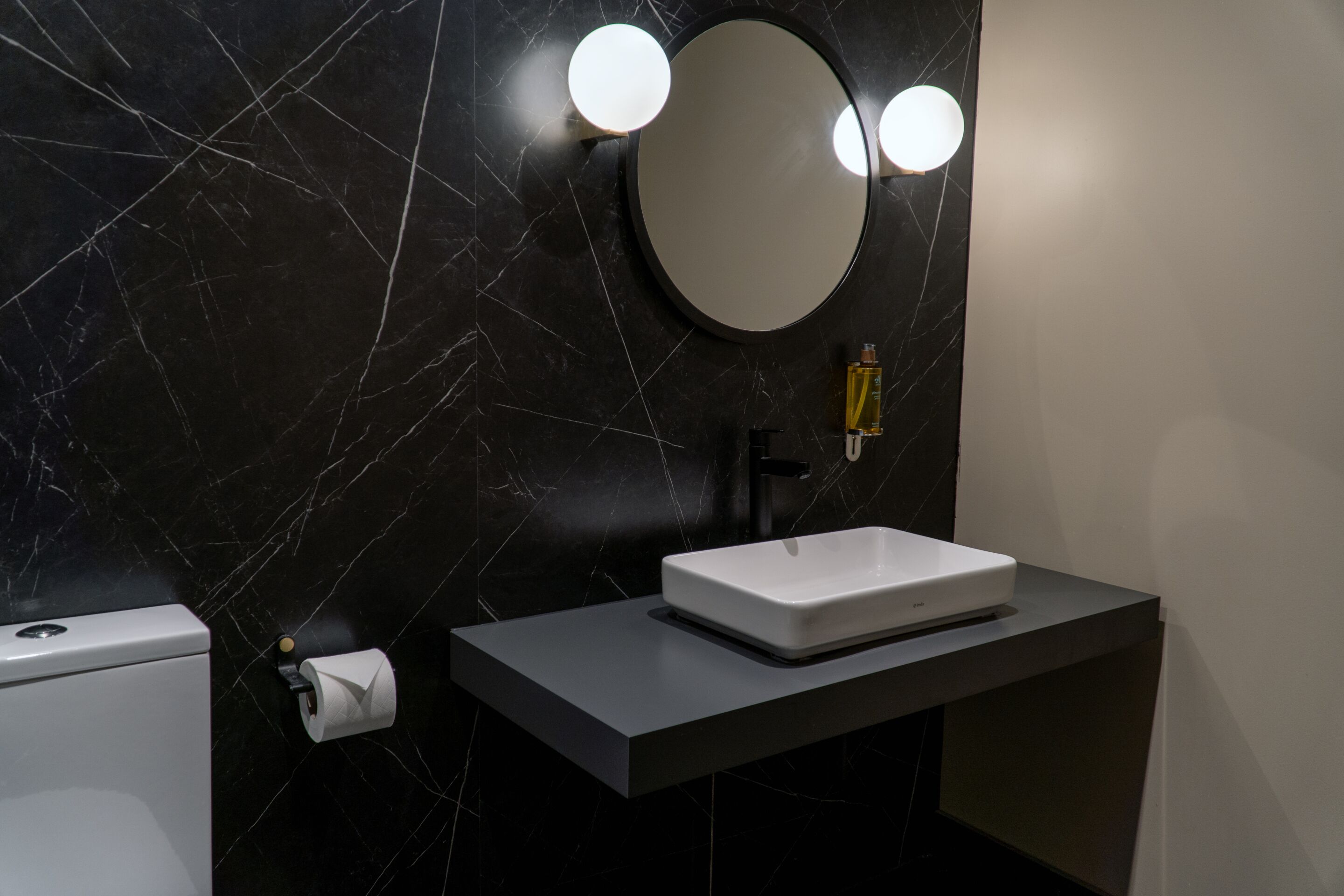 A modern bathroom with a black marble wall, a white sink, and a round mirror with two lights.