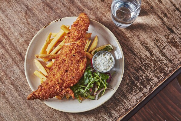 A plate of fish and chips with fries, a salad, and a small cup of sauce on a wooden table.