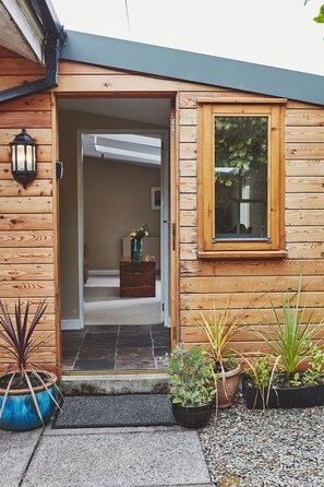 A wooden exterior with a door leading to an interior. There are potted plants outside and a small table with a vase inside.