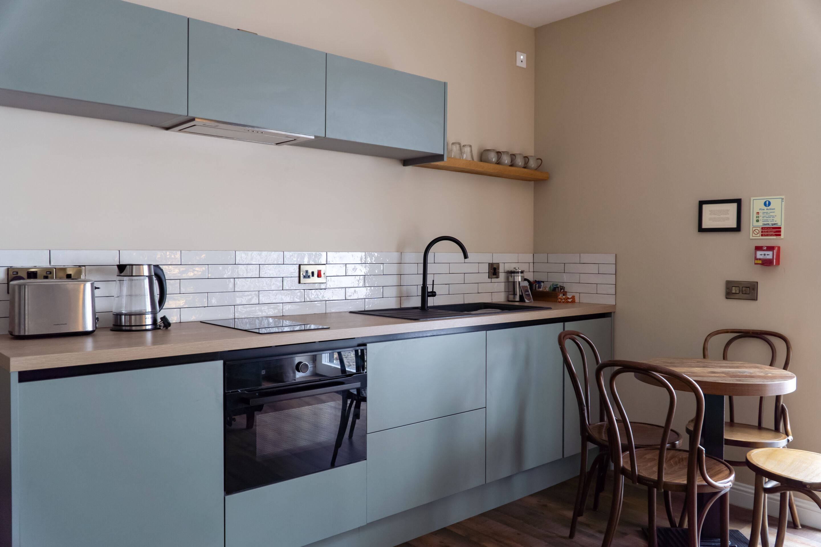 Eine moderne Küche mit hellblauen Schränken, einem Edelstahltoaster, einer Kaffeemaschine und einem Spülbecken. Es gibt Holzstühle und einen kleinen Tisch.