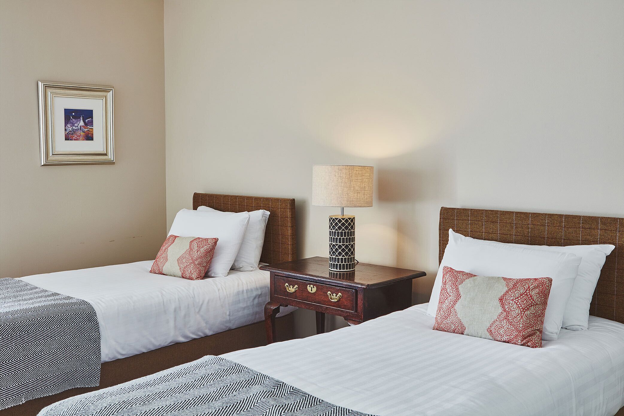 A hotel room with two single beds, a nightstand with a lamp, and a framed picture on the wall.