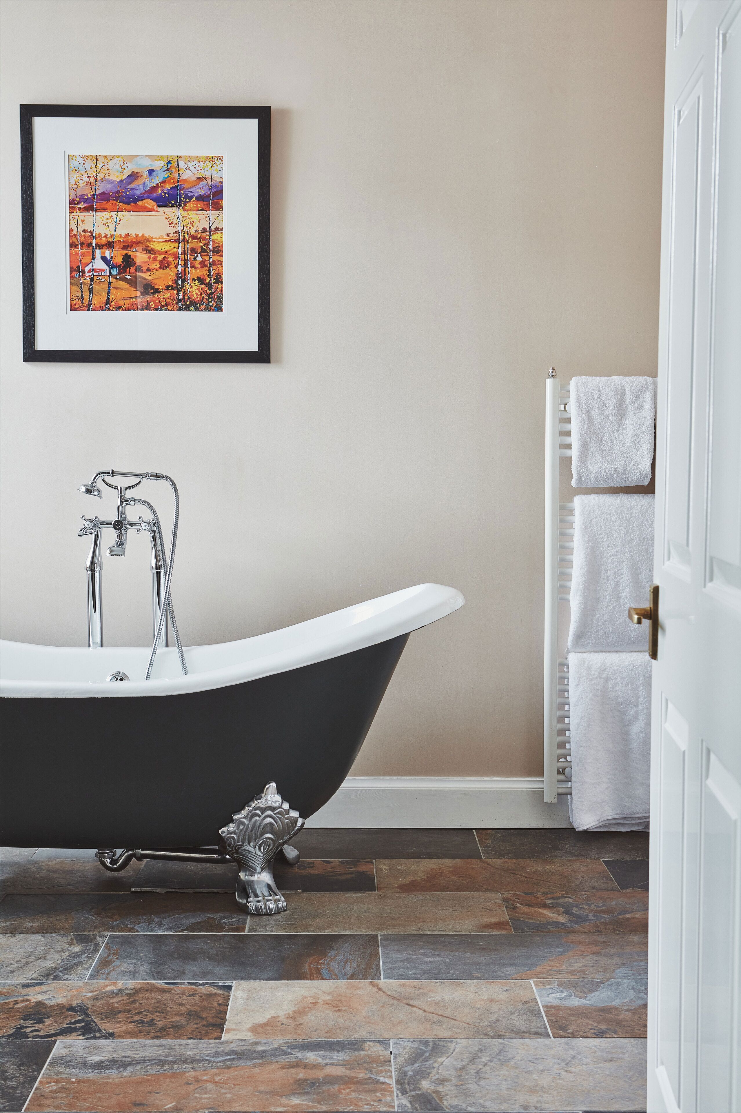 A bathroom with a freestanding bathtub, a framed landscape painting, and a towel rack with multiple towels.