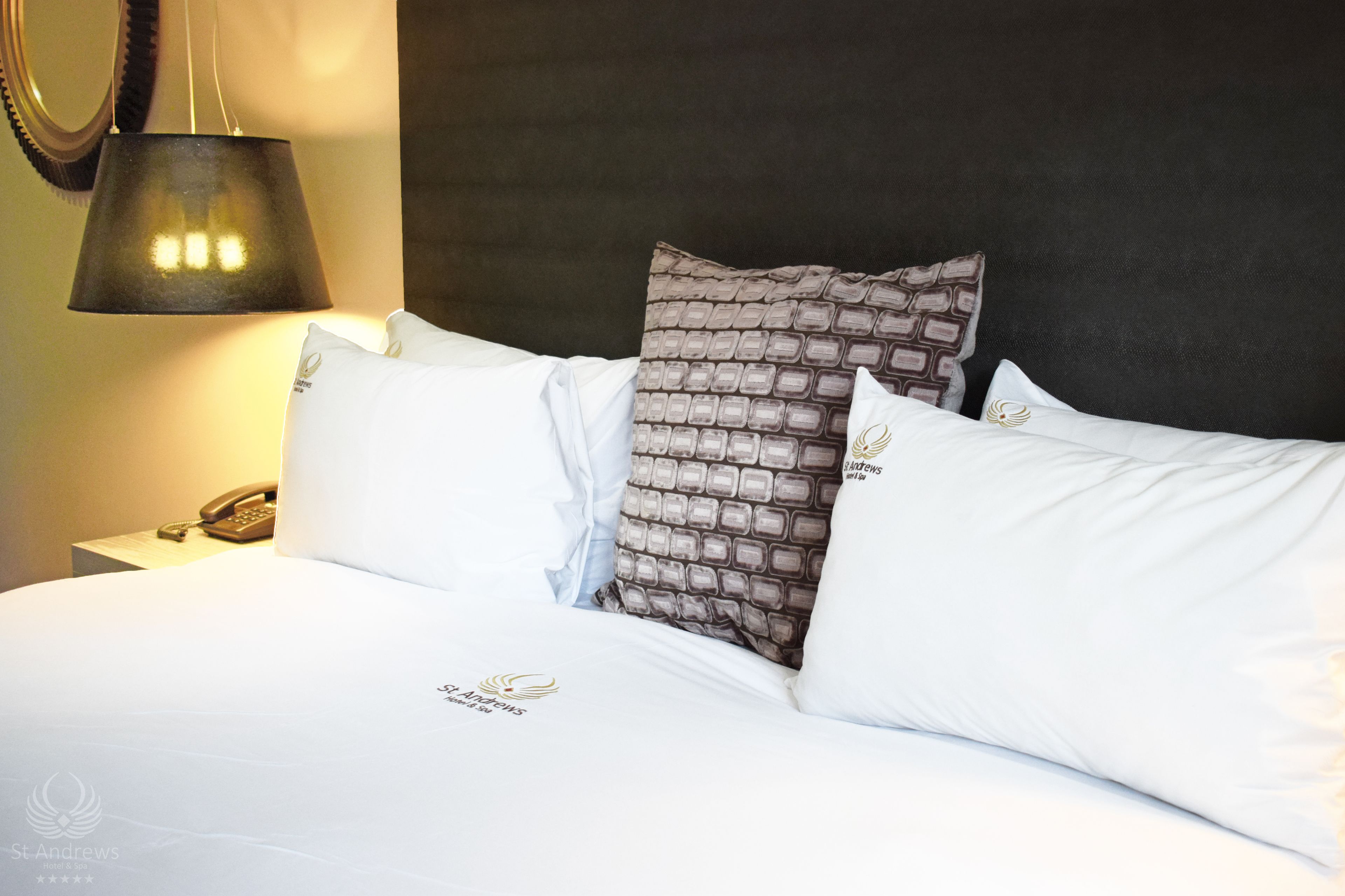 Una cama bien hecha con ropa de cama blanca y una almohada estampada, un teléfono en la mesita de noche y una lámpara sobre ella.