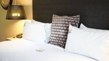 Una cama bien hecha con ropa de cama blanca y una almohada estampada, un teléfono en la mesita de noche y una lámpara sobre ella.