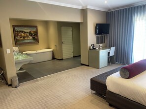 A hotel room with a bed, a desk, a TV, a painting, a bathtub, and a sink. - St Andrews Boutique Hotel (Germiston)
