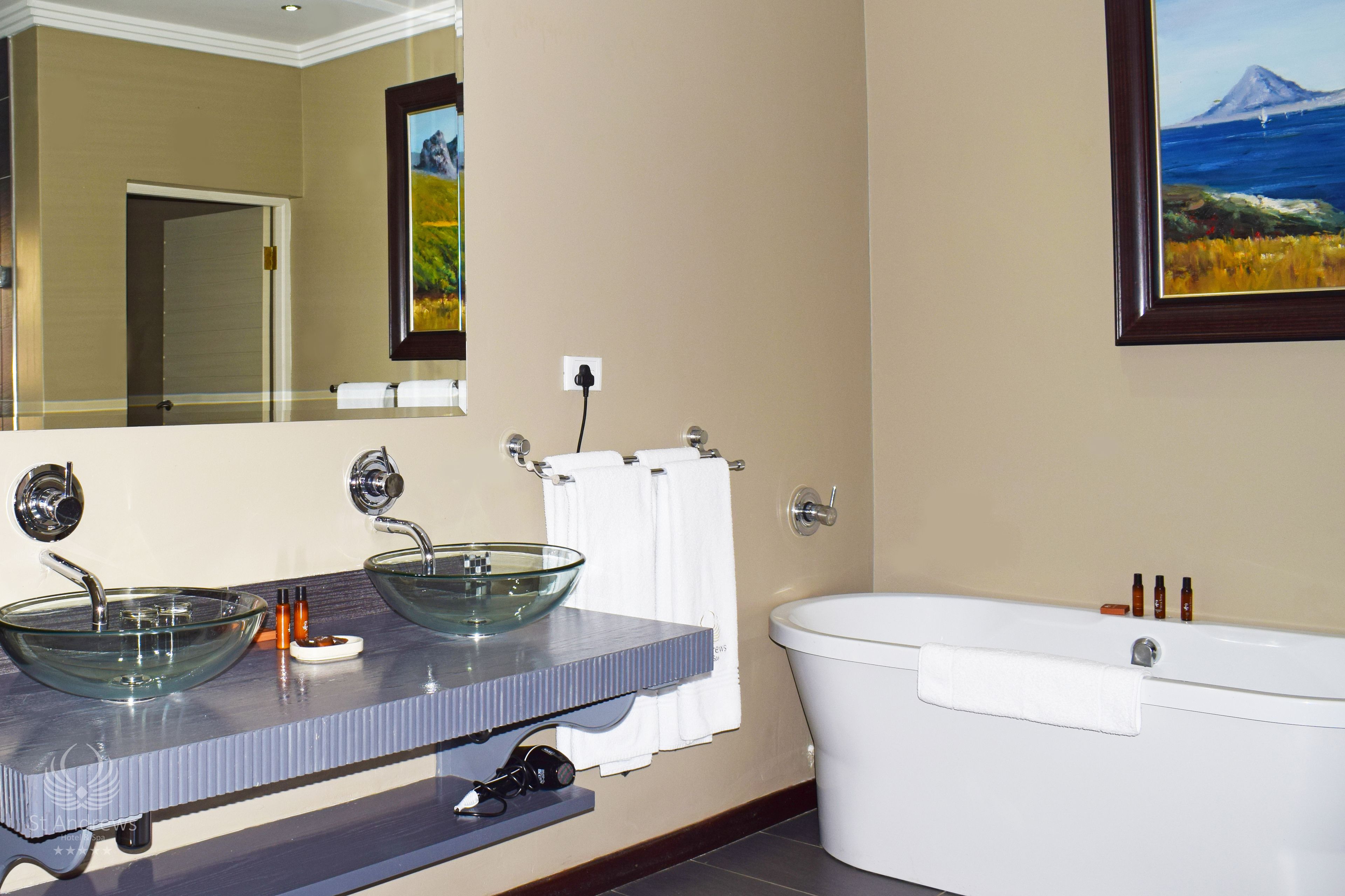 Ein Badezimmer mit einem Waschbecken aus Glas, einer weißen Badewanne und einem Spiegel, der eine Tür und ein Bild reflektiert.