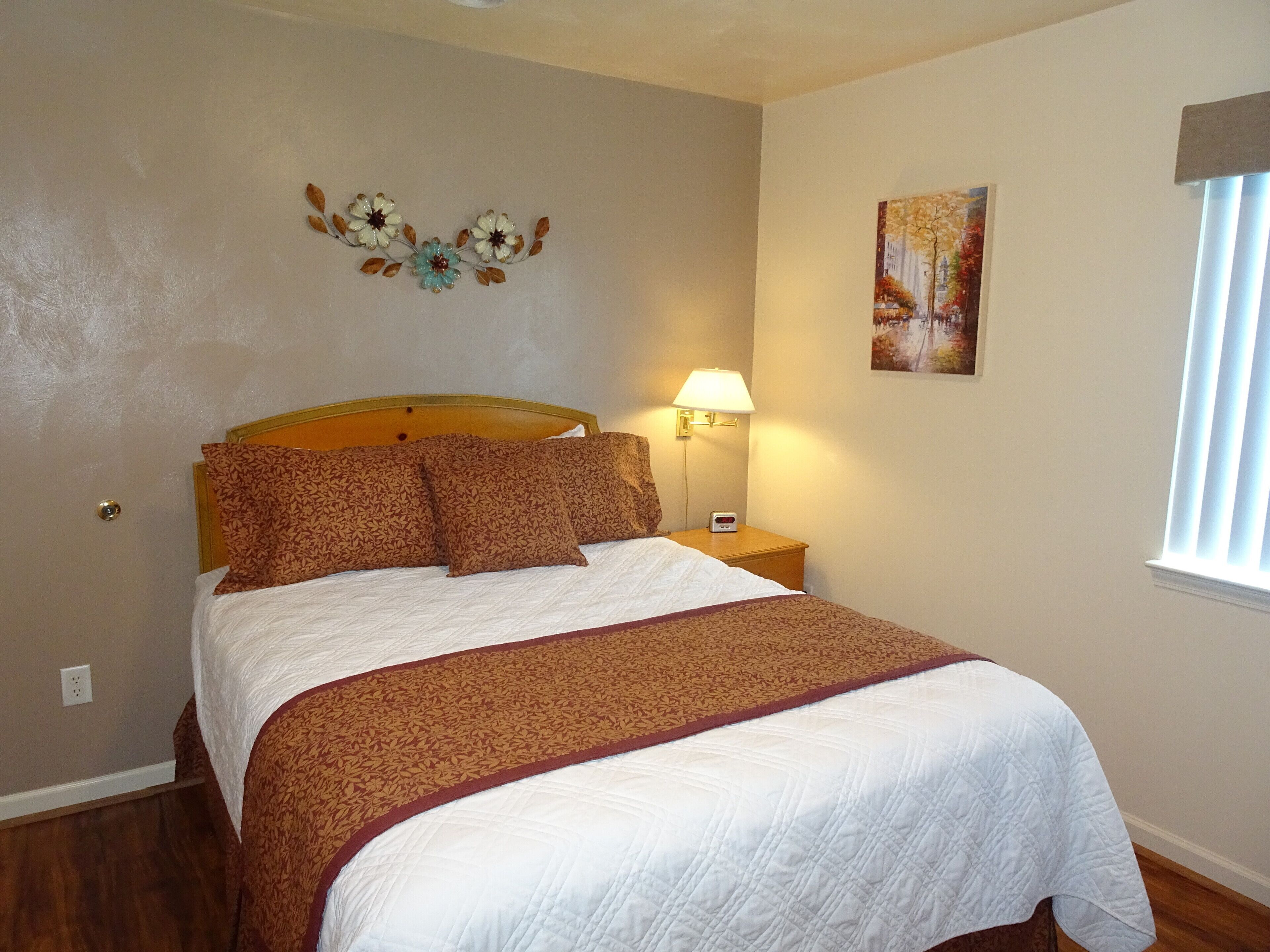 Een keurig opgemaakt bed met een bedsprei en kussens met een patroon, een aan de wand bevestigd decoratief bloemstuk, een bedlamp en een schilderij.