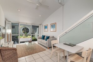A modern living room with a sofa, two chairs, a coffee table, and a dining table. There is a ceiling fan, a large window with curtains, and a glass door leading outside. - Caribbean Noosa (Noosa Heads)