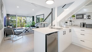 Una cocina moderna con un refrigerador empotrado, un microondas y una tetera. Hay un comedor con una mesa y sillas, y una escalera que lleva a otro nivel.