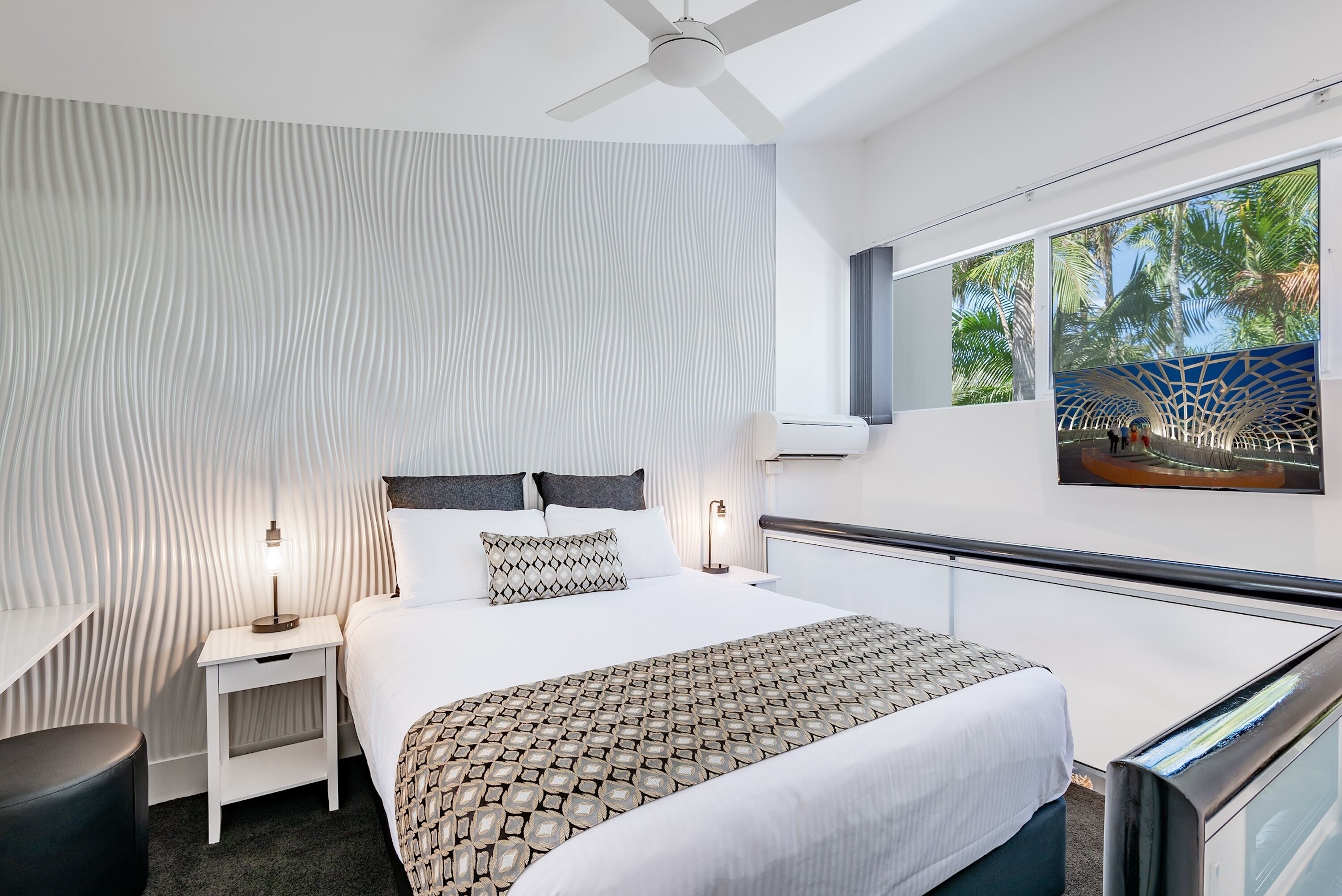 A modern hotel room with a large bed, a nightstand, a desk, and a view of a tropical outdoor area.