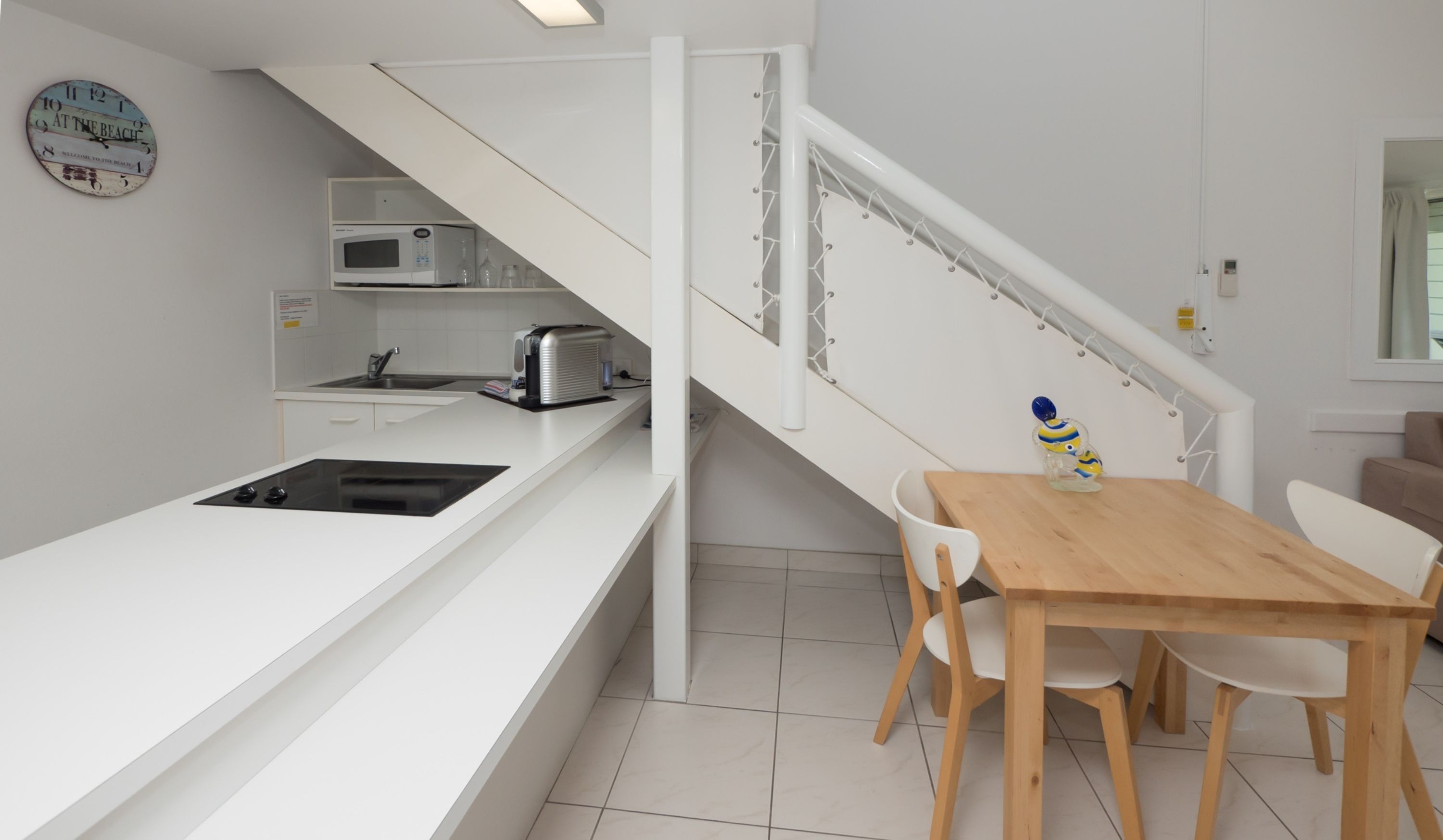 A modern kitchen with a white countertop, a built-in microwave, and a toaster. There is a wooden dining table with chairs and a decorative glass object on it. A staircase with a white railing leads to another level.