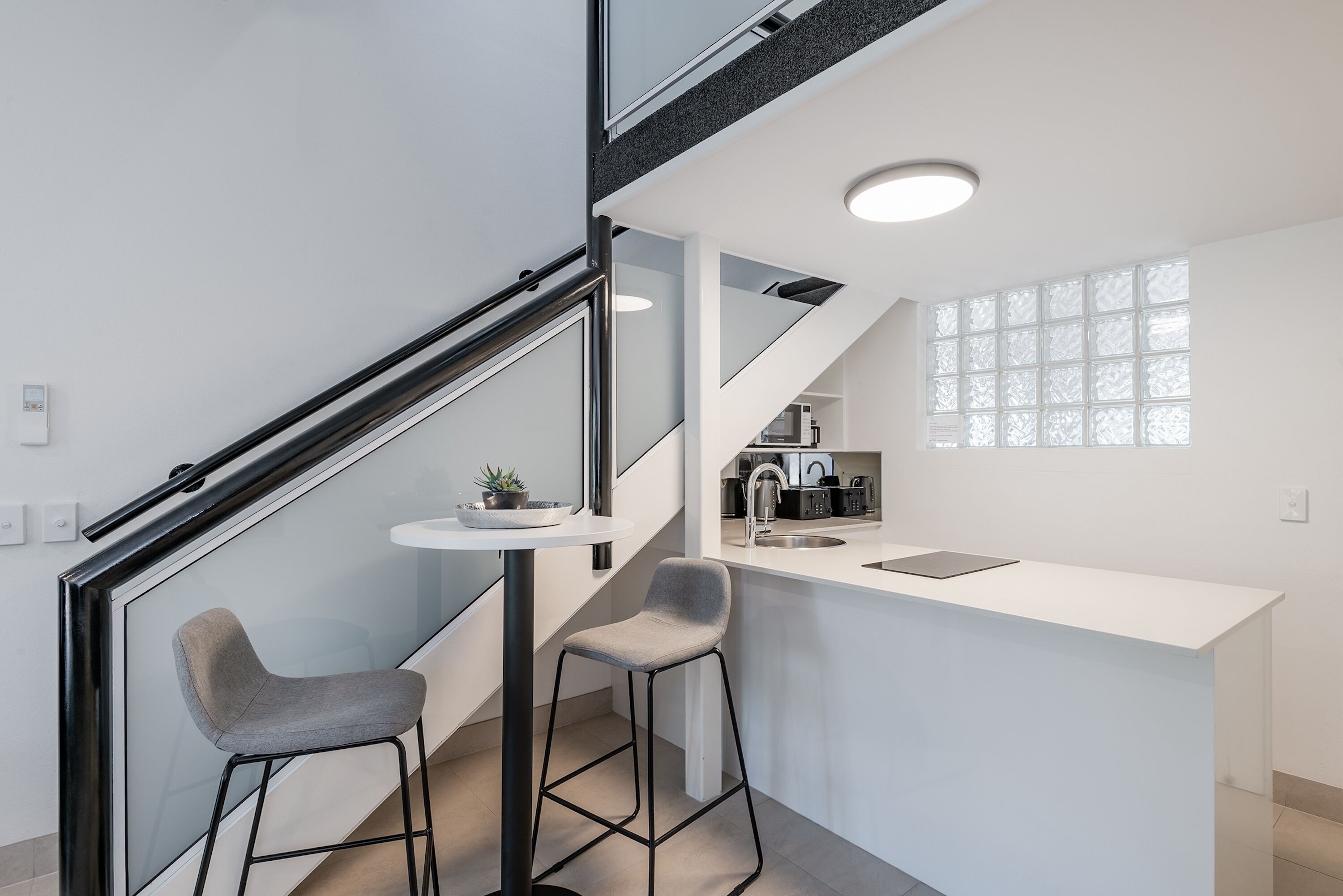 A modern kitchen with a white countertop, two bar stools, a small round table, and a staircase with glass railings.