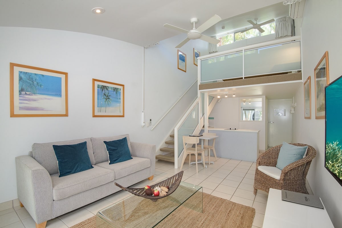 A modern living room with a sofa, a wicker chair, a glass coffee table, and a staircase leading to an upper level.