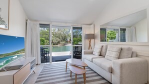 Una sala moderna con sofá, una mesa de centro y televisor. Cuenta con una puerta corrediza de vidrio que da a un espacio exterior con piscina y vegetación.