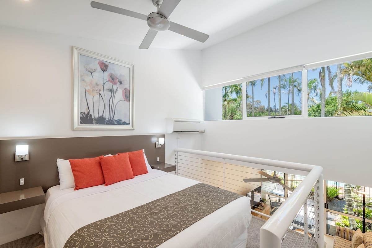 A modern hotel room with a large bed, a ceiling fan, a painting, and a view of the outdoors.