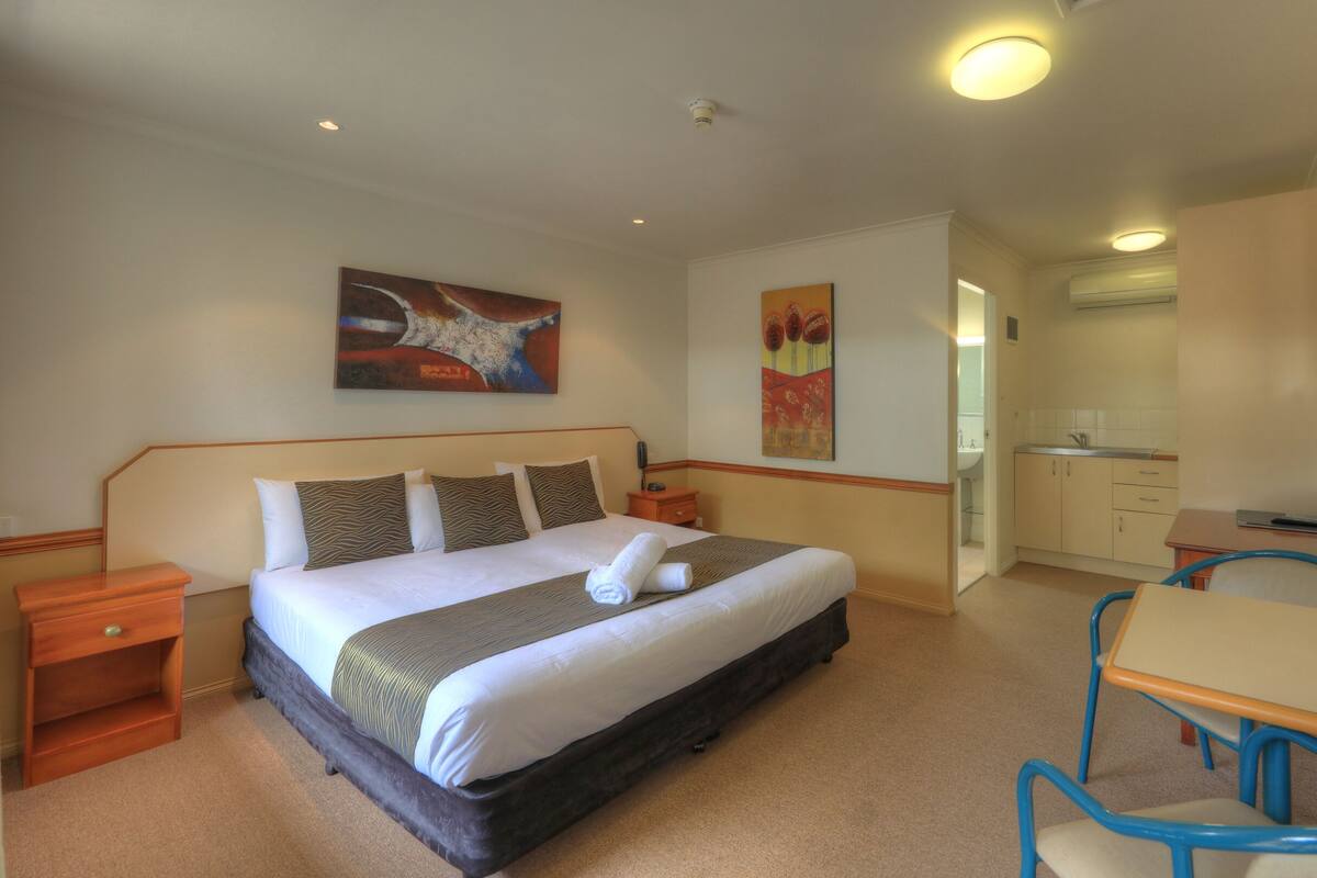 A hotel room with a large bed, a desk, a chair, a small table, and a kitchenette.