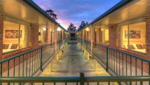 A corridor with brick walls and glass windows, leading to a covered area with a view of trees. - Bent Street Motor Inn (South Grafton)