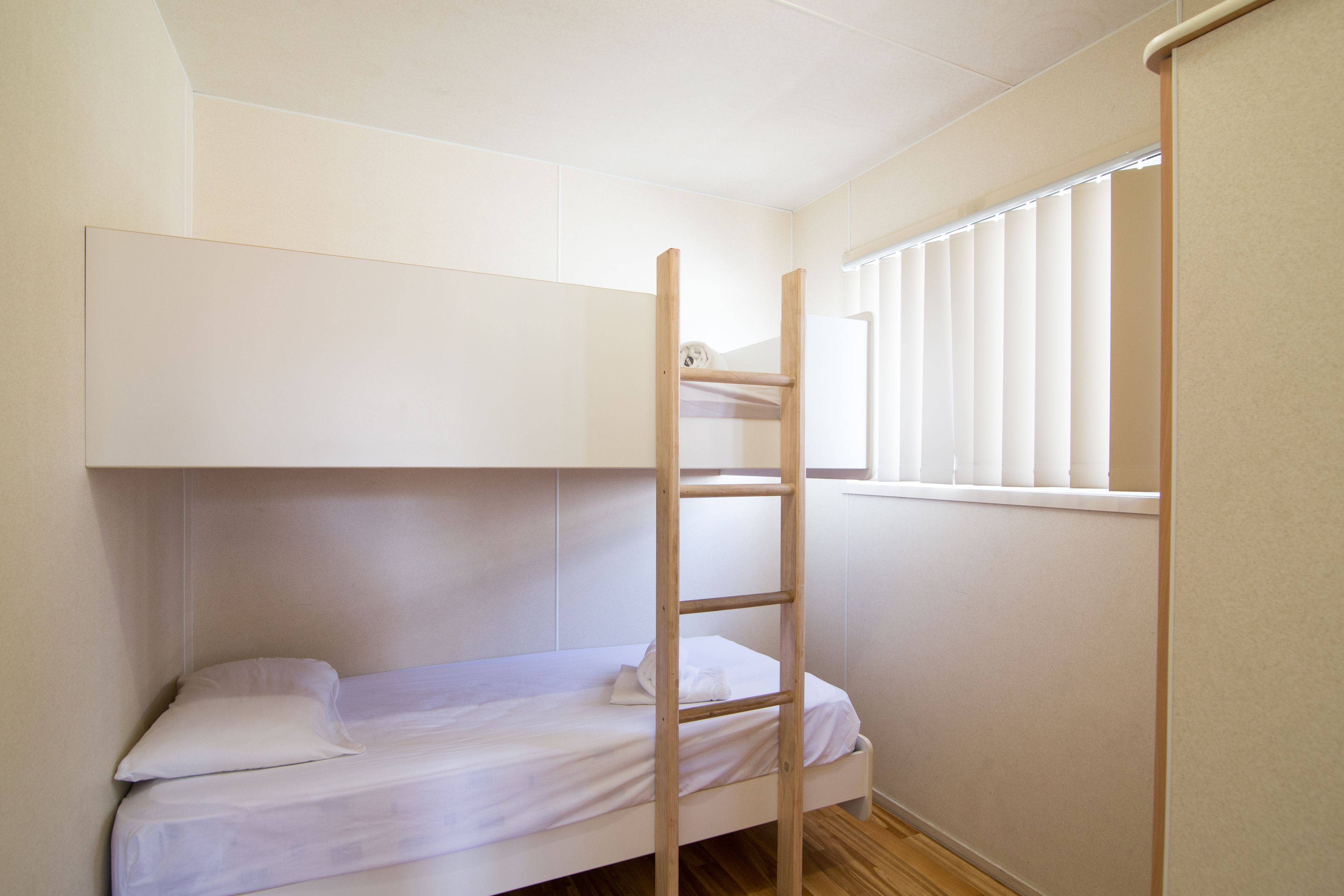 A bunk bed with a wooden ladder, a window with blinds, and a plain wall.