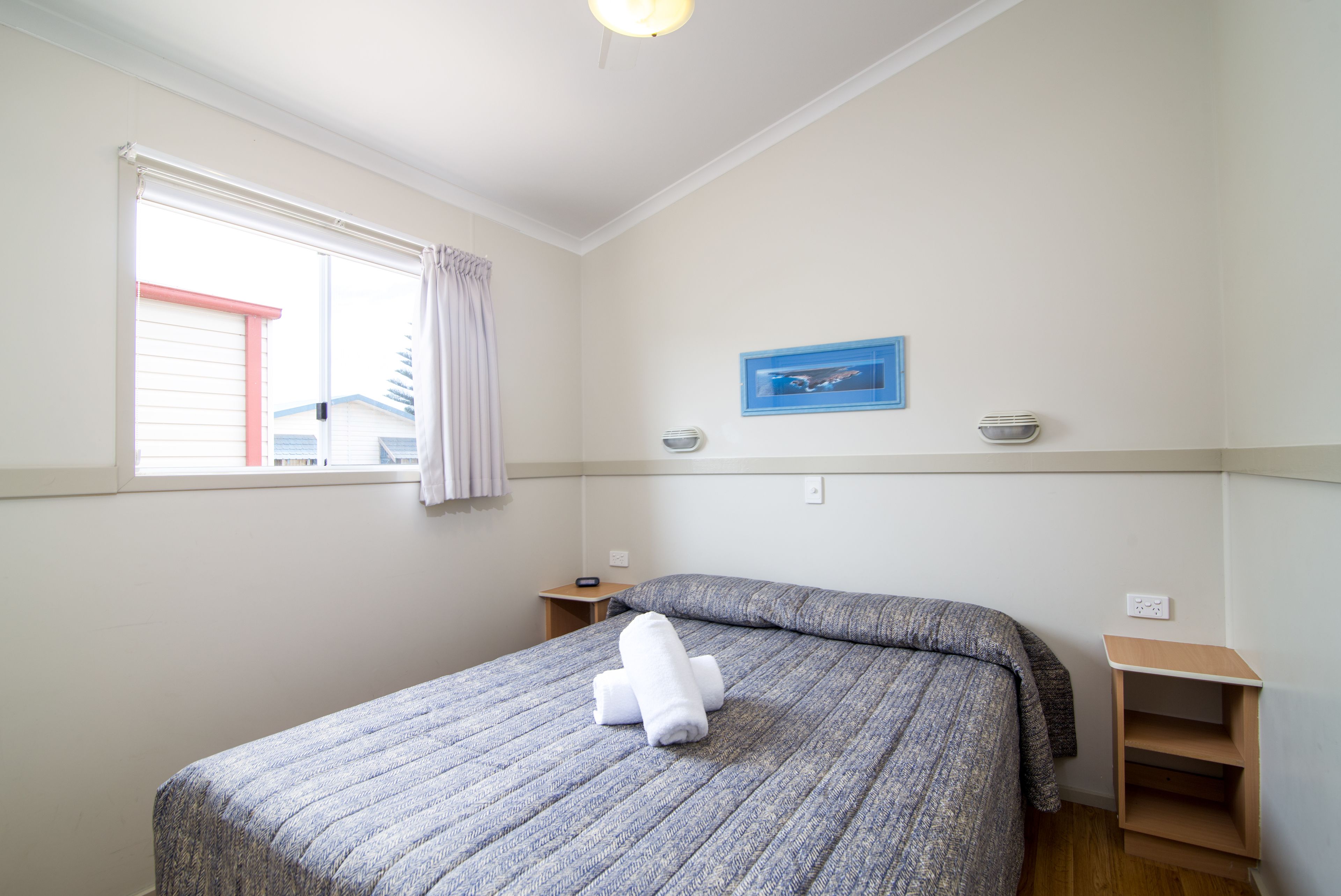 A bedroom with a bed, a window with curtains, a bedside table, and a wall-mounted shelf.