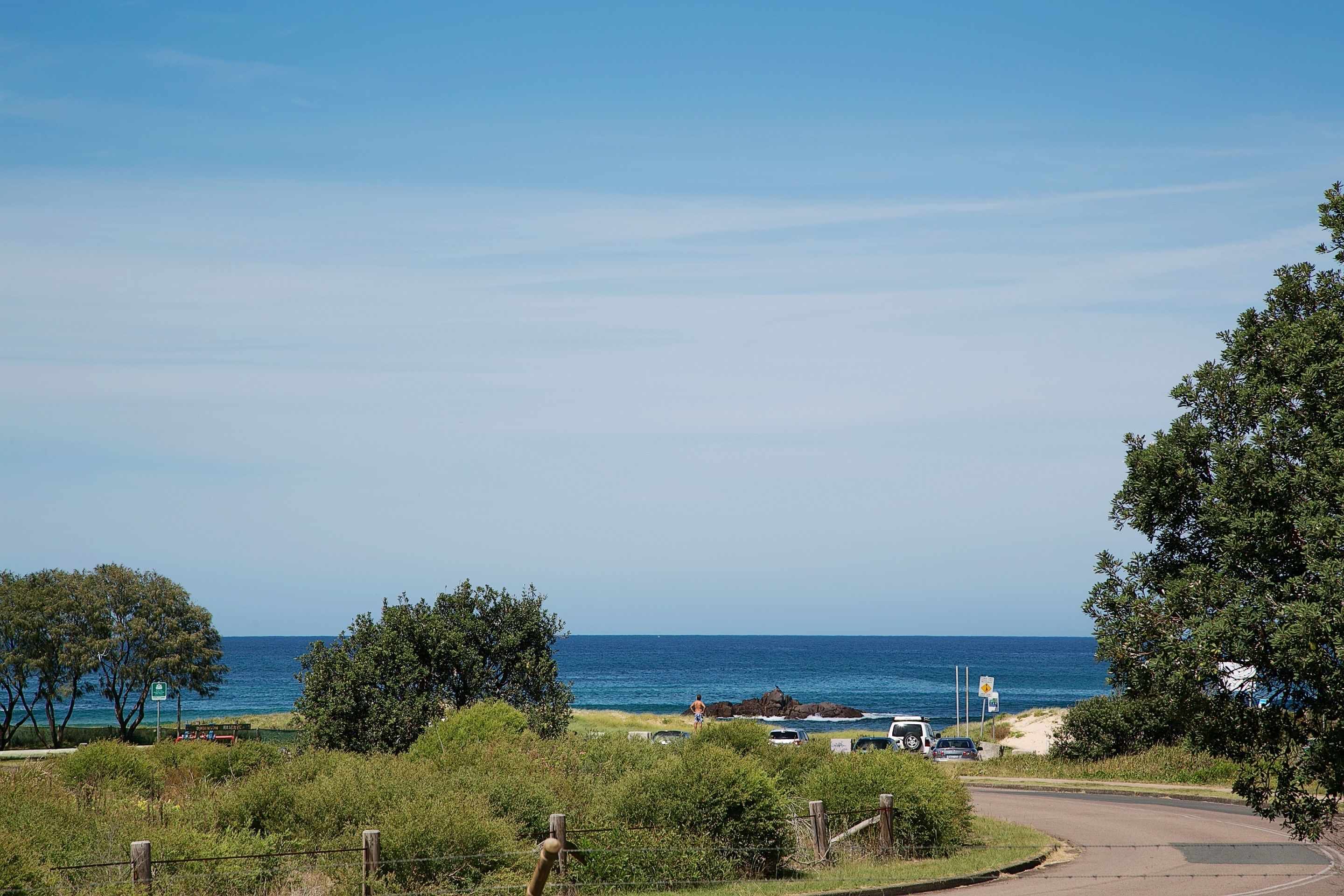 A coastal road with a view of the ocean, a beach, and a few parked cars.