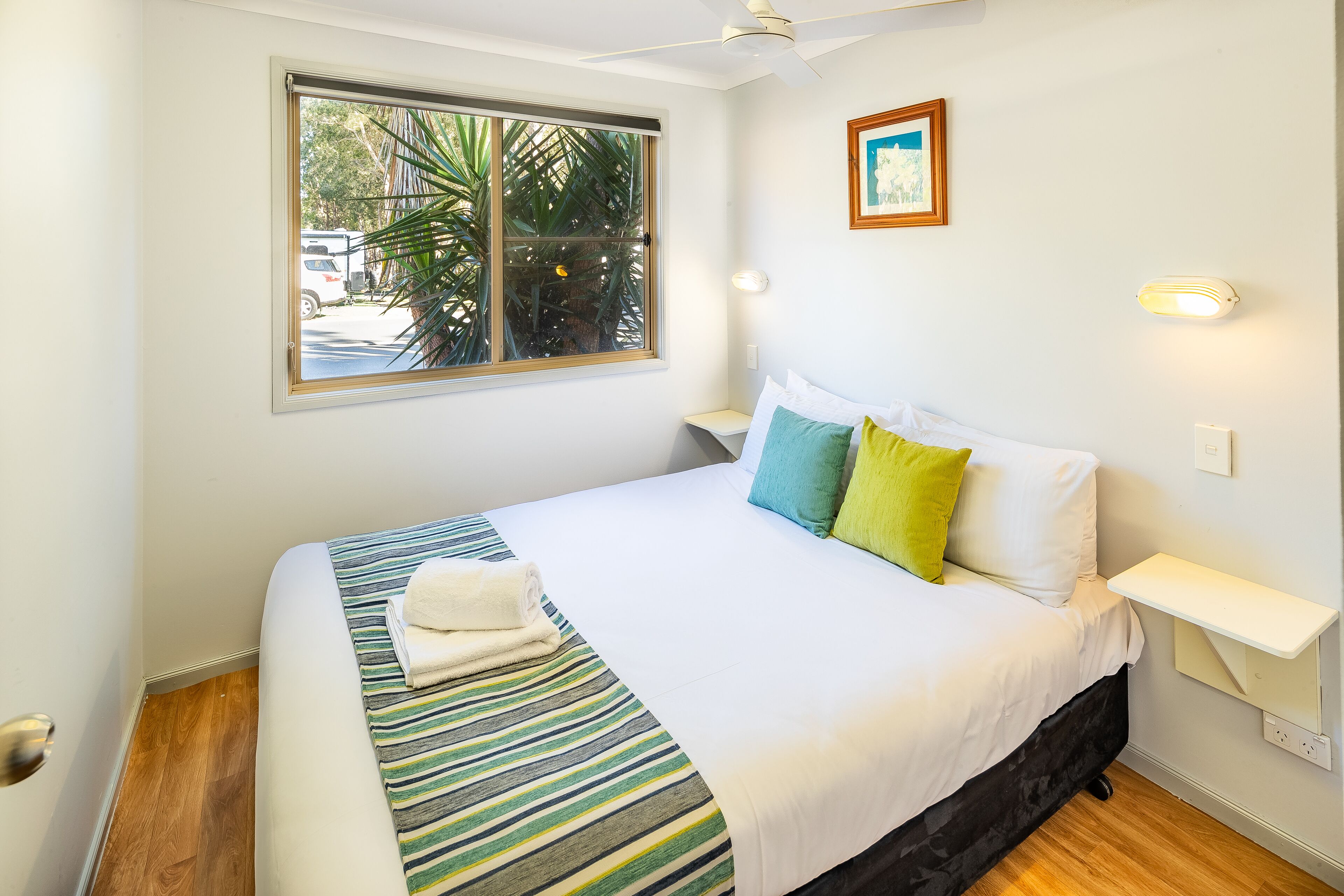 A neatly made bed with a striped bedspread, pillows, and a folded towel. There is a window with a view of palm trees and a parked car outside.