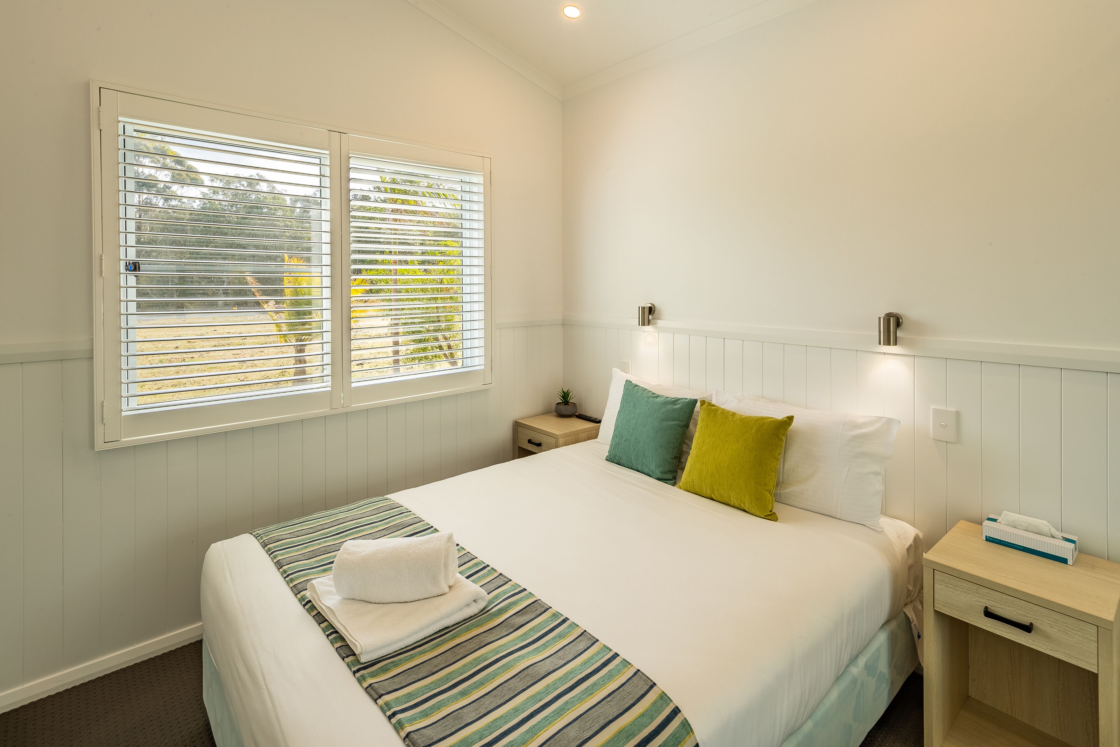 A neatly made bed with a striped bedspread, two pillows, and a bedside table with a lamp and a tissue box.