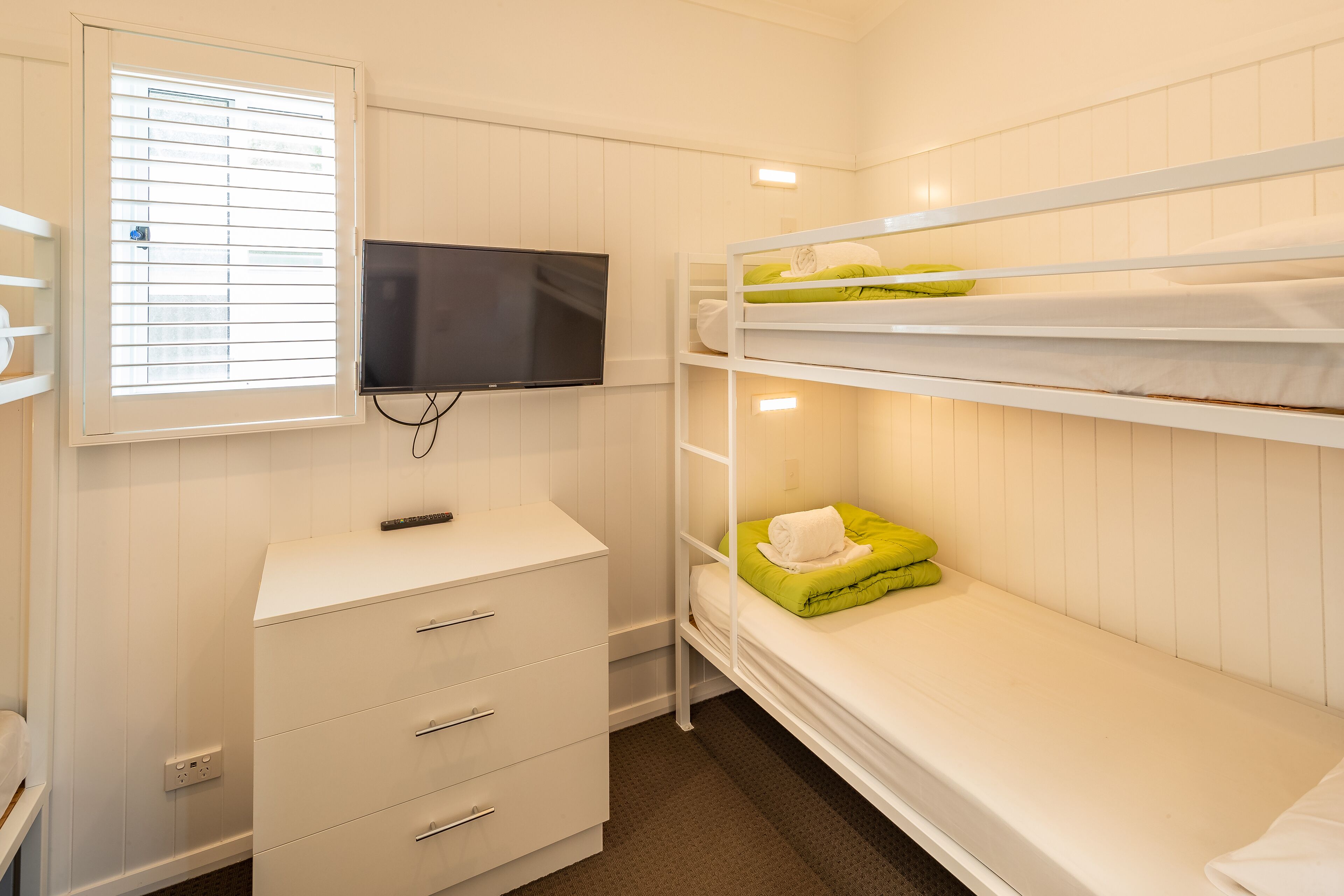 A bunk bed room with a TV mounted on the wall, a dresser, and a window with blinds.