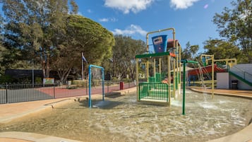 Un terrain de jeux doté d’une zone ludique avec des jeux d’eau, entouré d’arbres et délimité par une clôture.