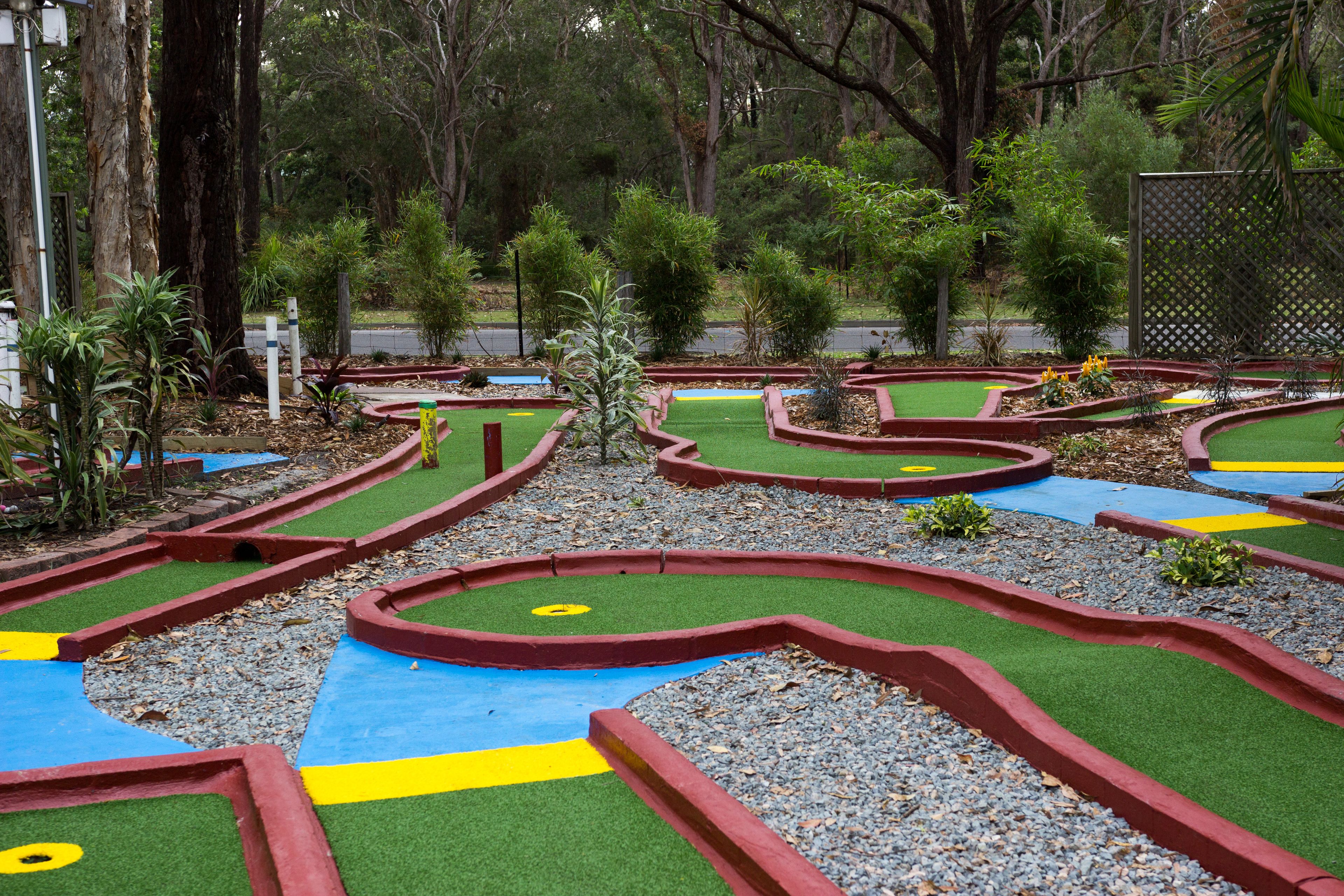 Un parcours de mini-golf avec des tapis de jeu verts et bleus, entouré d’arbres et de plantes.