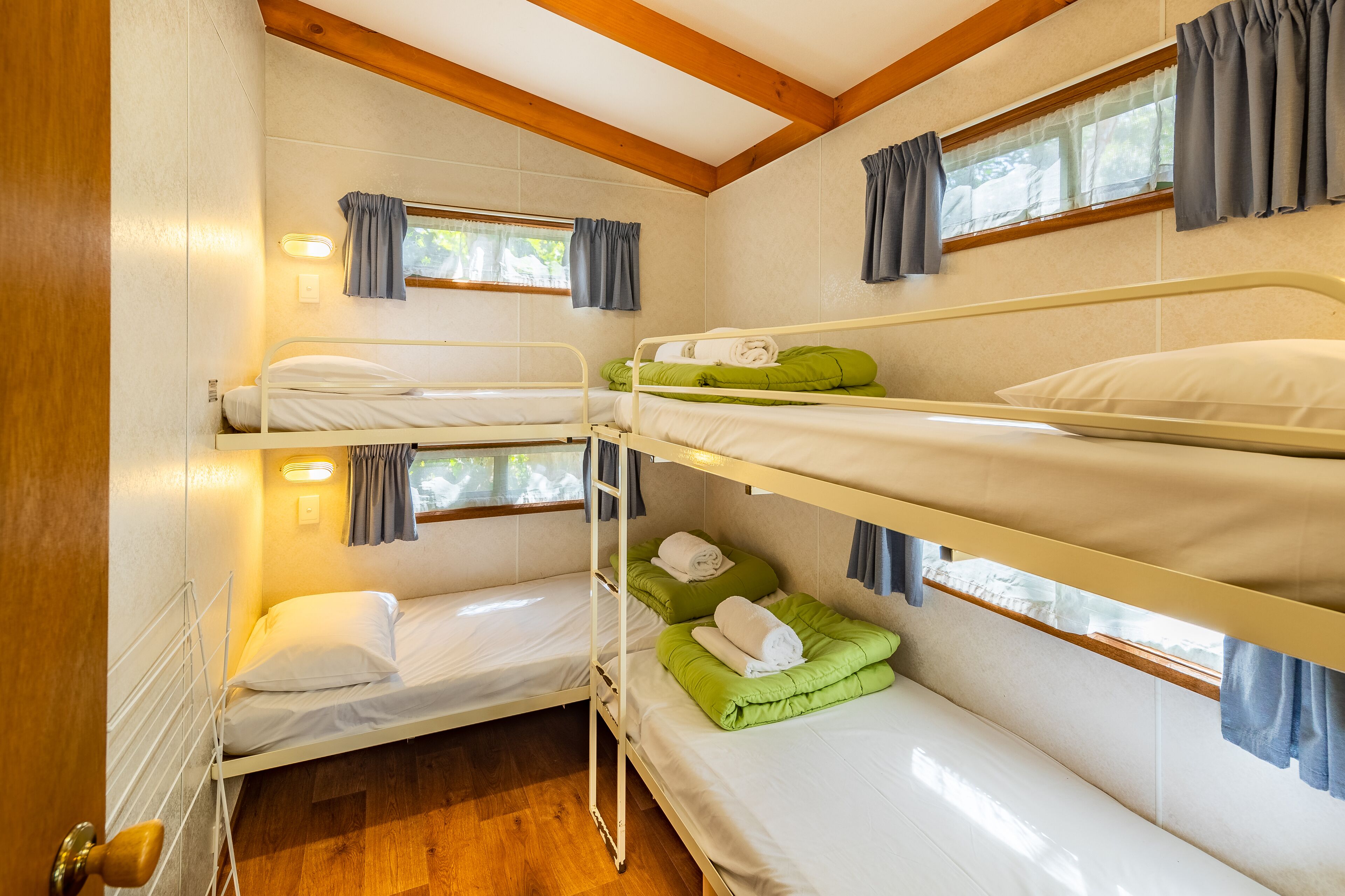A bunk bed room with three levels, wooden beams on the ceiling, and windows with curtains.
