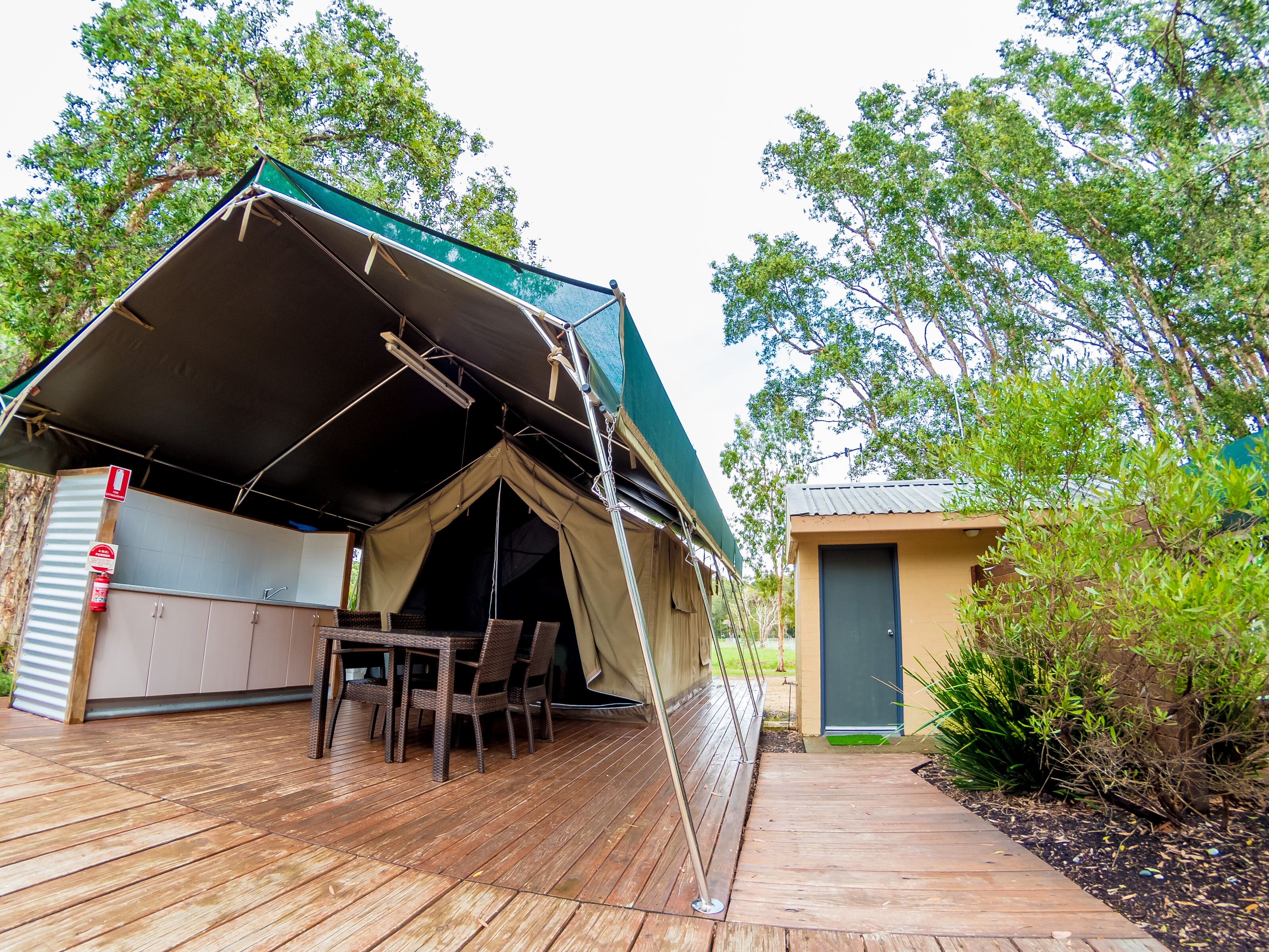 A tented outdoor area with a table and chairs, a small building, and a pathway.