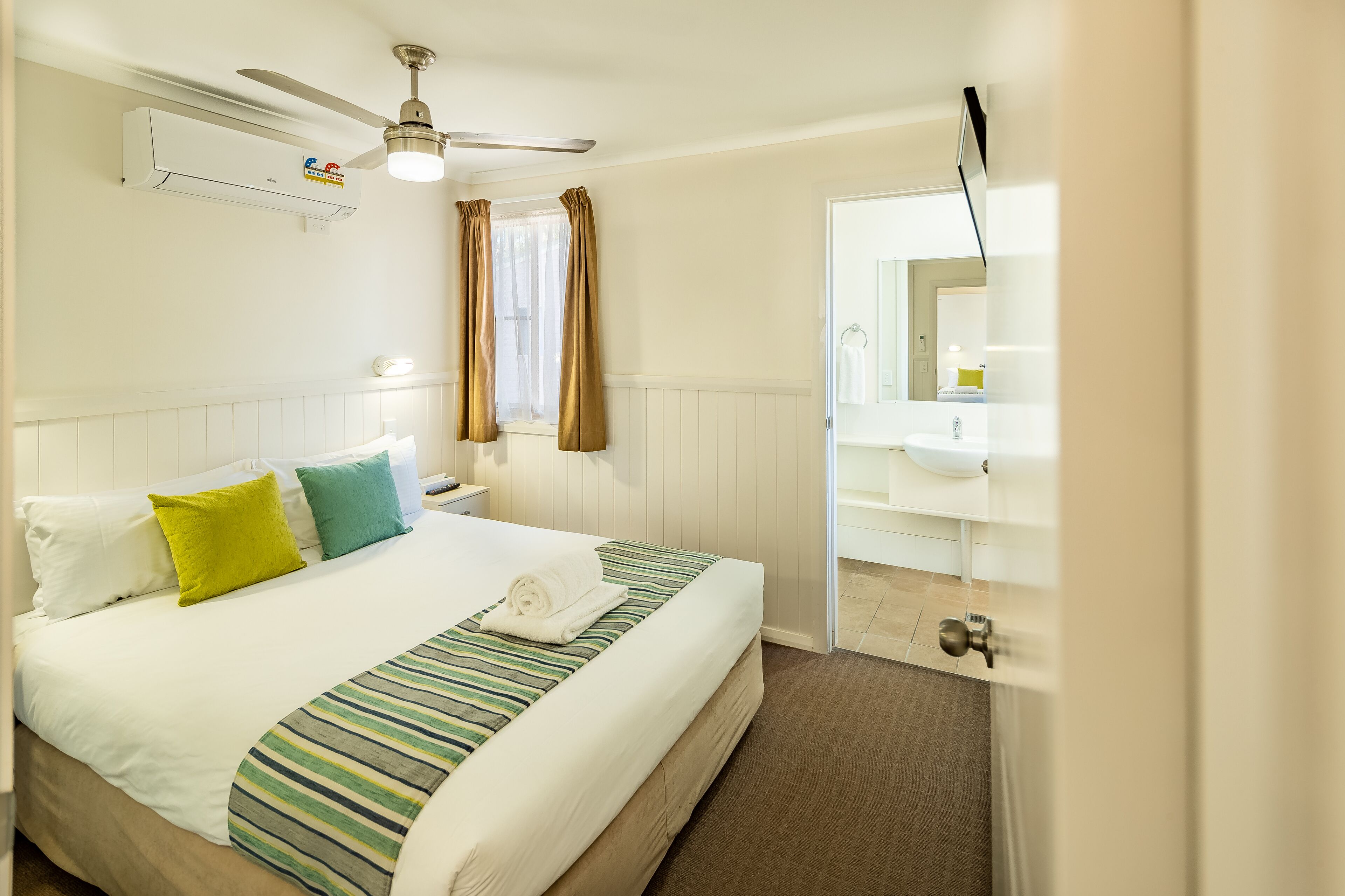 A hotel room with a bed, a ceiling fan, a window with curtains, a bathroom with a sink and mirror.