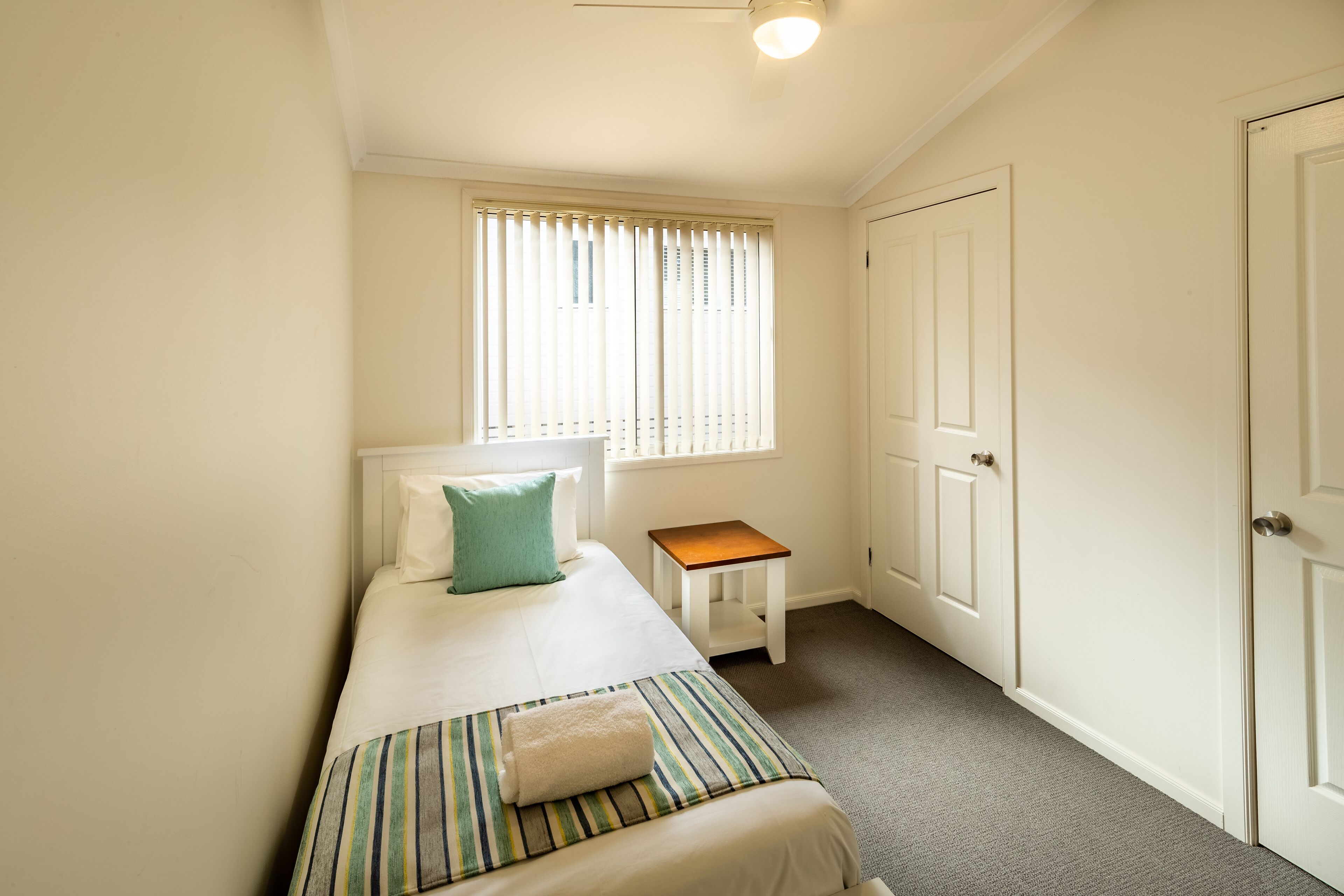 A small, clean room with a single bed, a nightstand, a window with blinds, and a door.