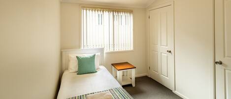 A small, clean room with a single bed, a nightstand, a window with blinds, and a door.