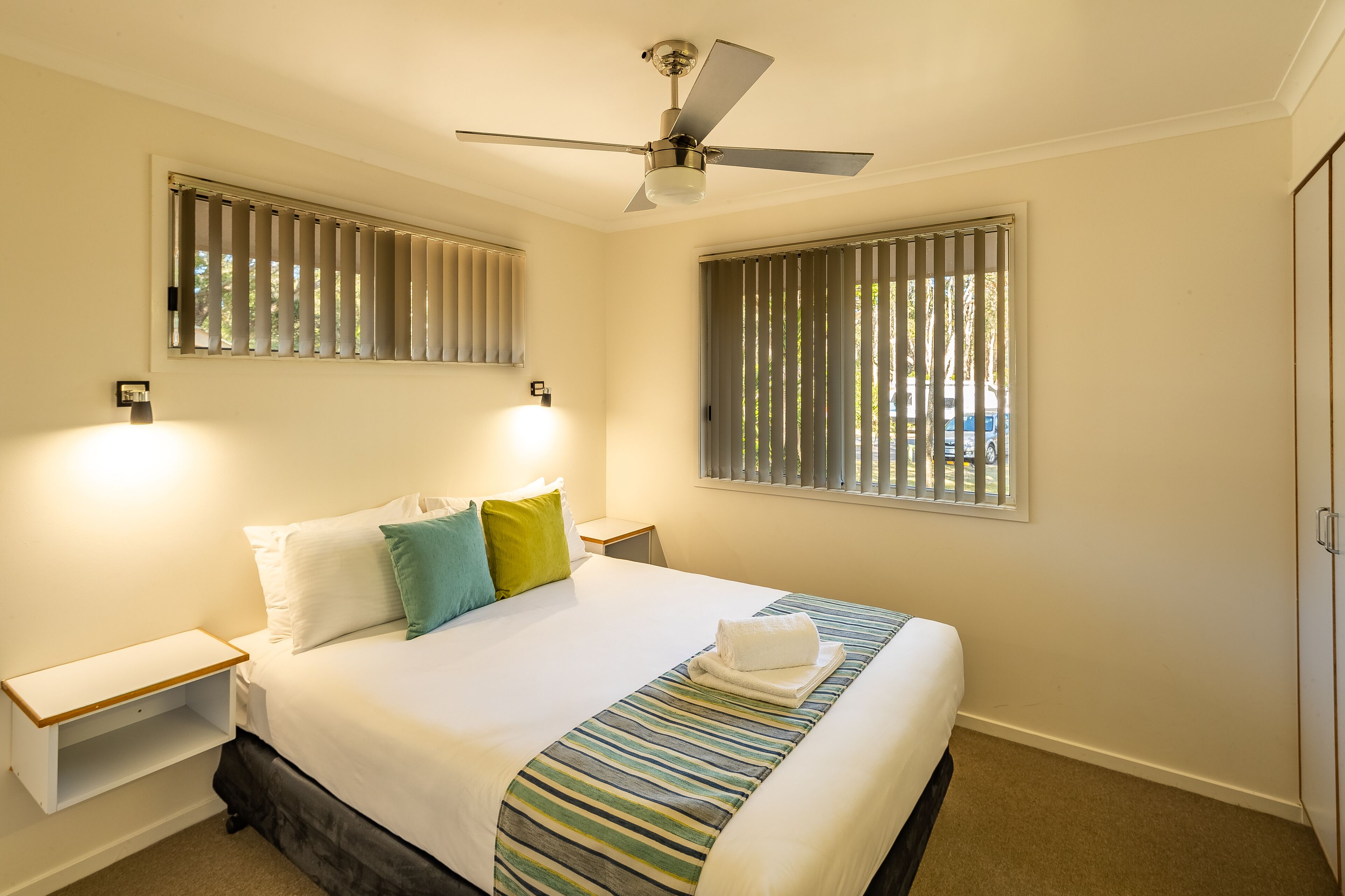 A neatly arranged hotel room with a bed, a nightstand, a bedside table, a ceiling fan, and two windows with blinds.