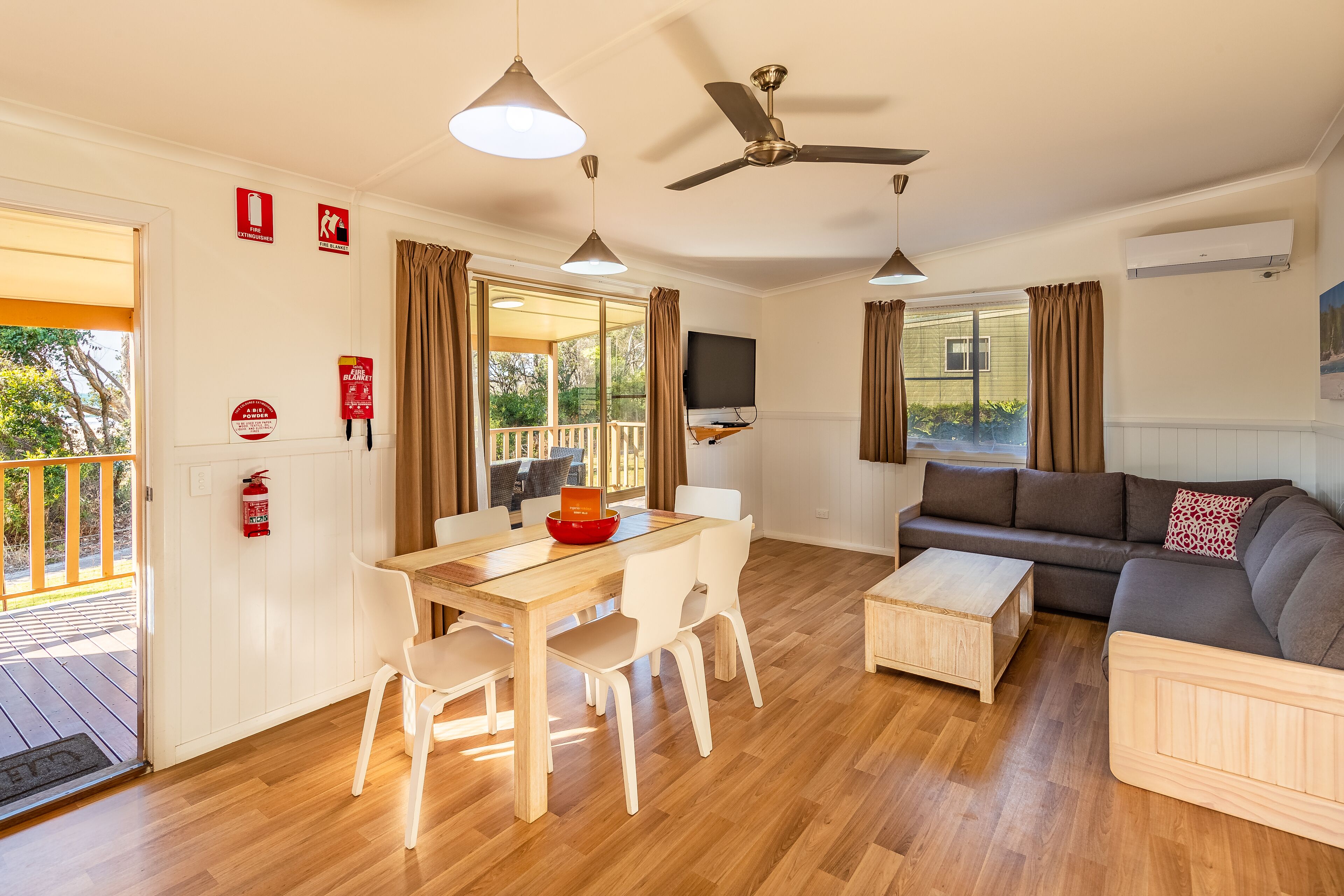 Un vaste espace de vie comprenant une table à manger en bois, des chaises blanches, un canapé gris et un ventilateur de plafond.