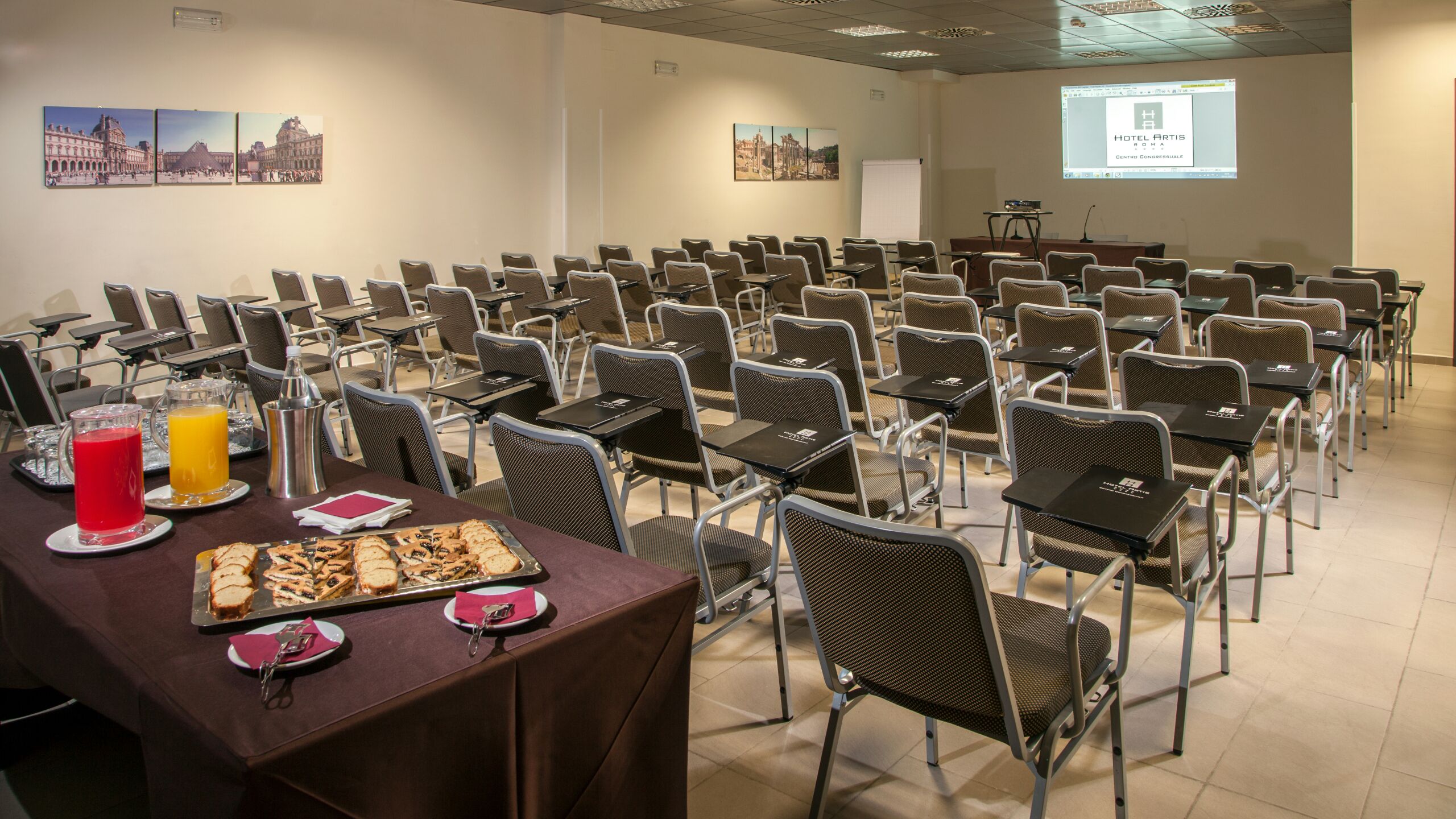 Une salle de conférence avec des rangées de chaises et de tables, un écran de projection affichant le nom « HÔTEL ARTIS », et une table de buffet garnie de plats et de boissons.