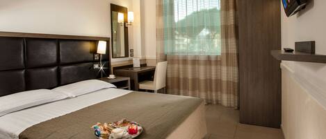 A hotel room with a bed, a desk, a chair, a tray with breakfast items, a TV, and a window with curtains.