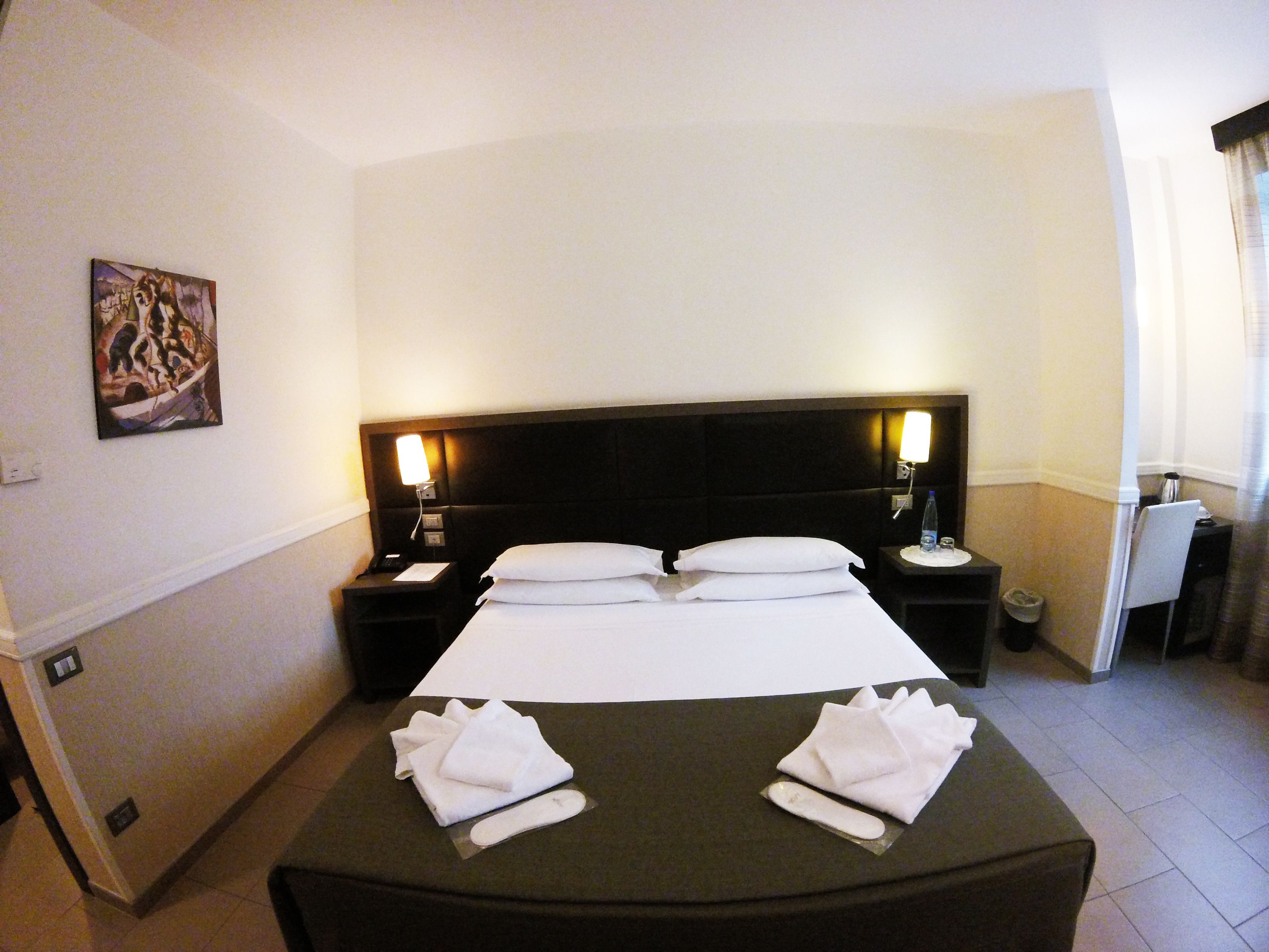 A hotel room with a large bed, two bedside tables with lamps, a painting on the wall, and a small desk with a chair.