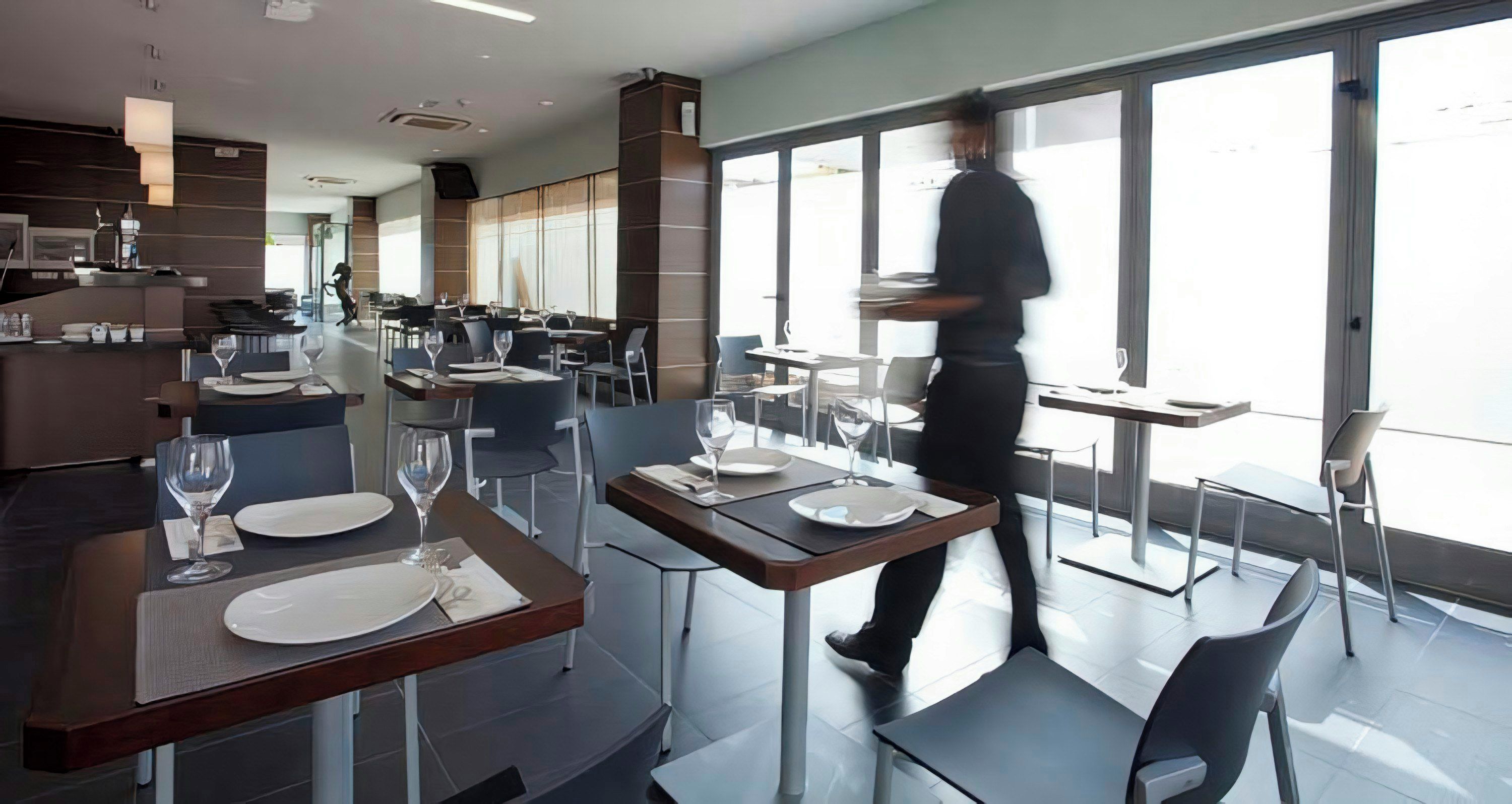 Interior moderno de restaurante con mesas dispuestas para comer, un camarero en movimiento y grandes ventanales que dejan entrar luz natural.