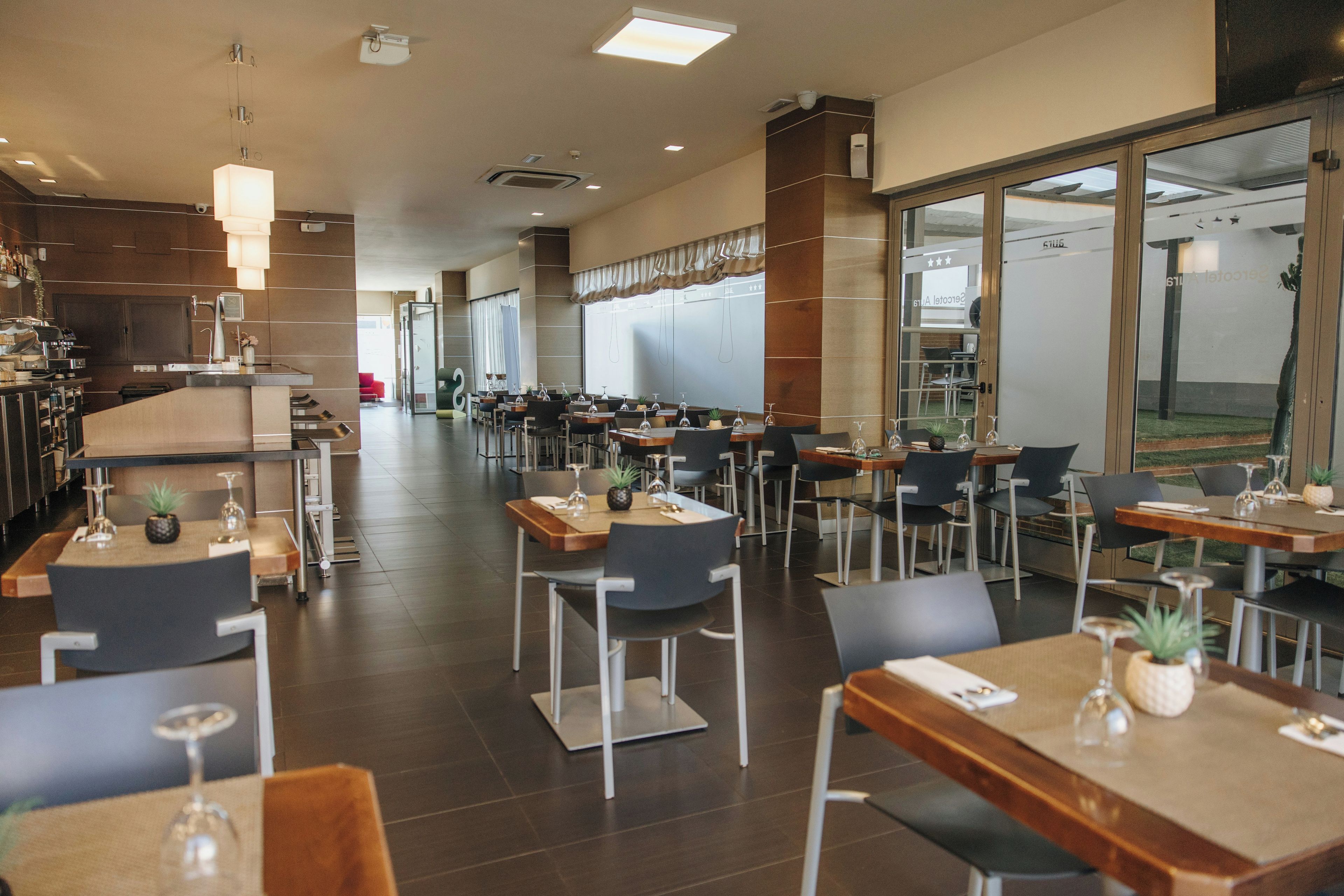 Un restaurant modern amb diverses taules i cadirees, una zona de cuina i grans finestres.