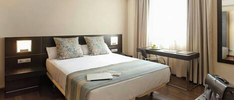 Habitación de hotel con cama, mesitas de noche, escritorio, silla, maleta y un amplio ventanal con cortinas.