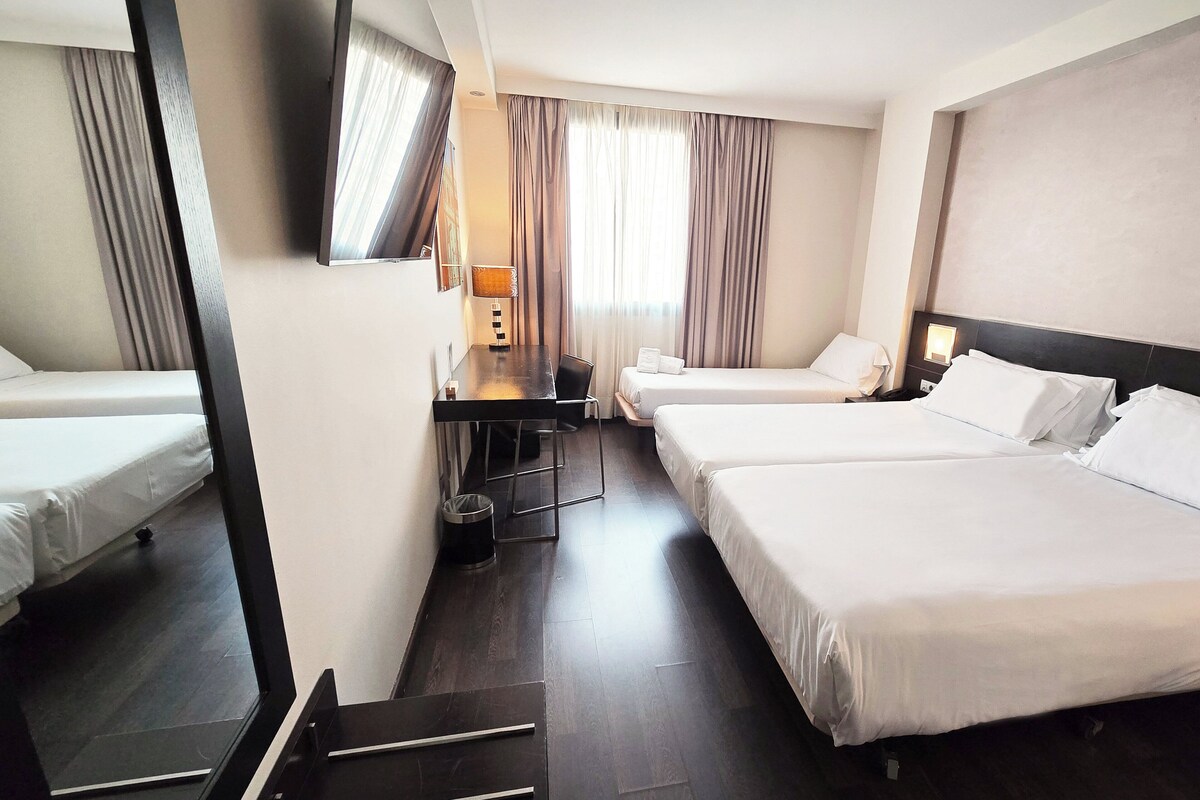 A hotel room with two beds, a desk, a chair, a TV, and a window with curtains.