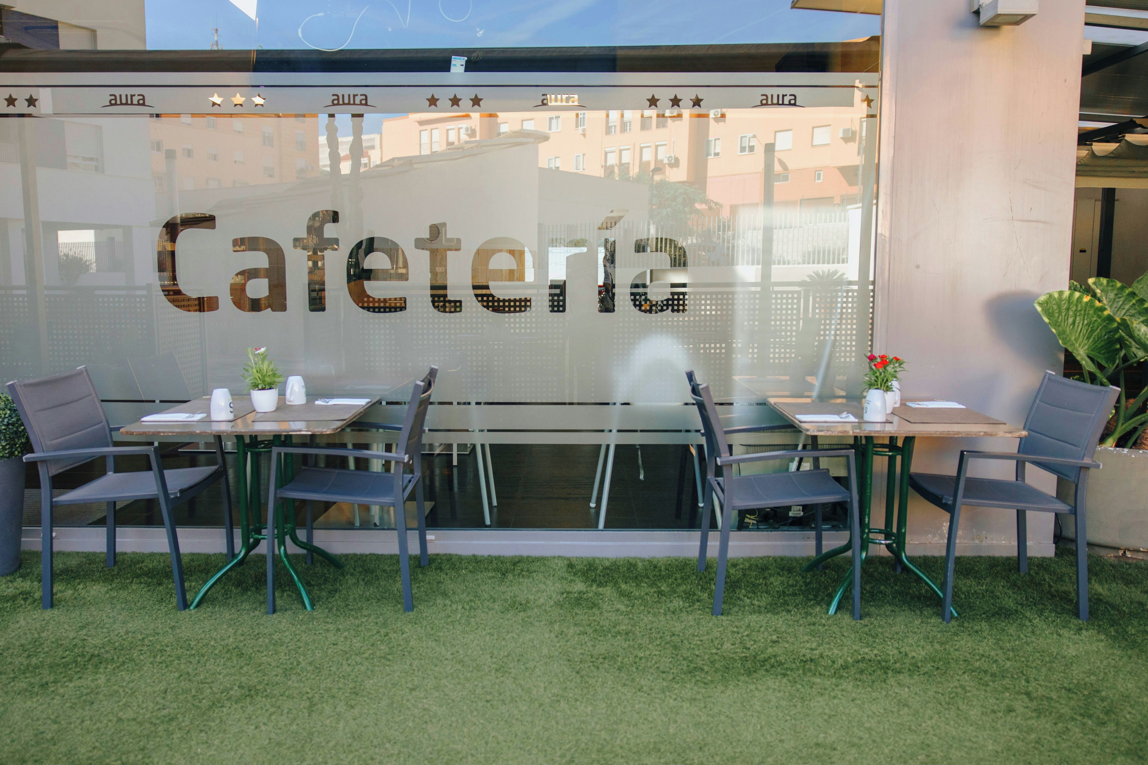 Un área de estar al aire libre con mesas y sillas, una partición de vidrio con la palabra 'Cafeteria' y el reflejo de edificios al fondo.