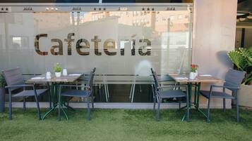 Un área de estar al aire libre con mesas y sillas, una partición de vidrio con la palabra 'Cafeteria' y el reflejo de edificios al fondo.