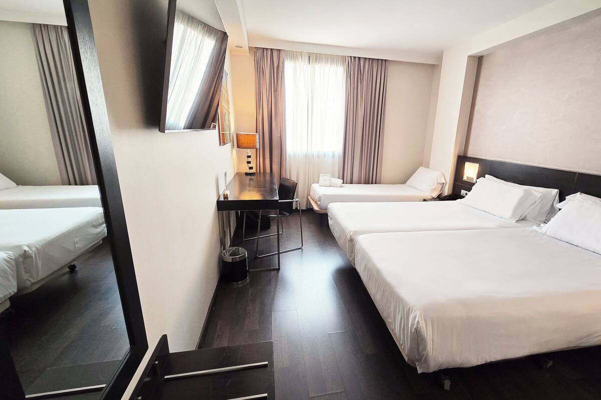 A hotel room with two beds, a desk, a chair, a TV, and a window with curtains.