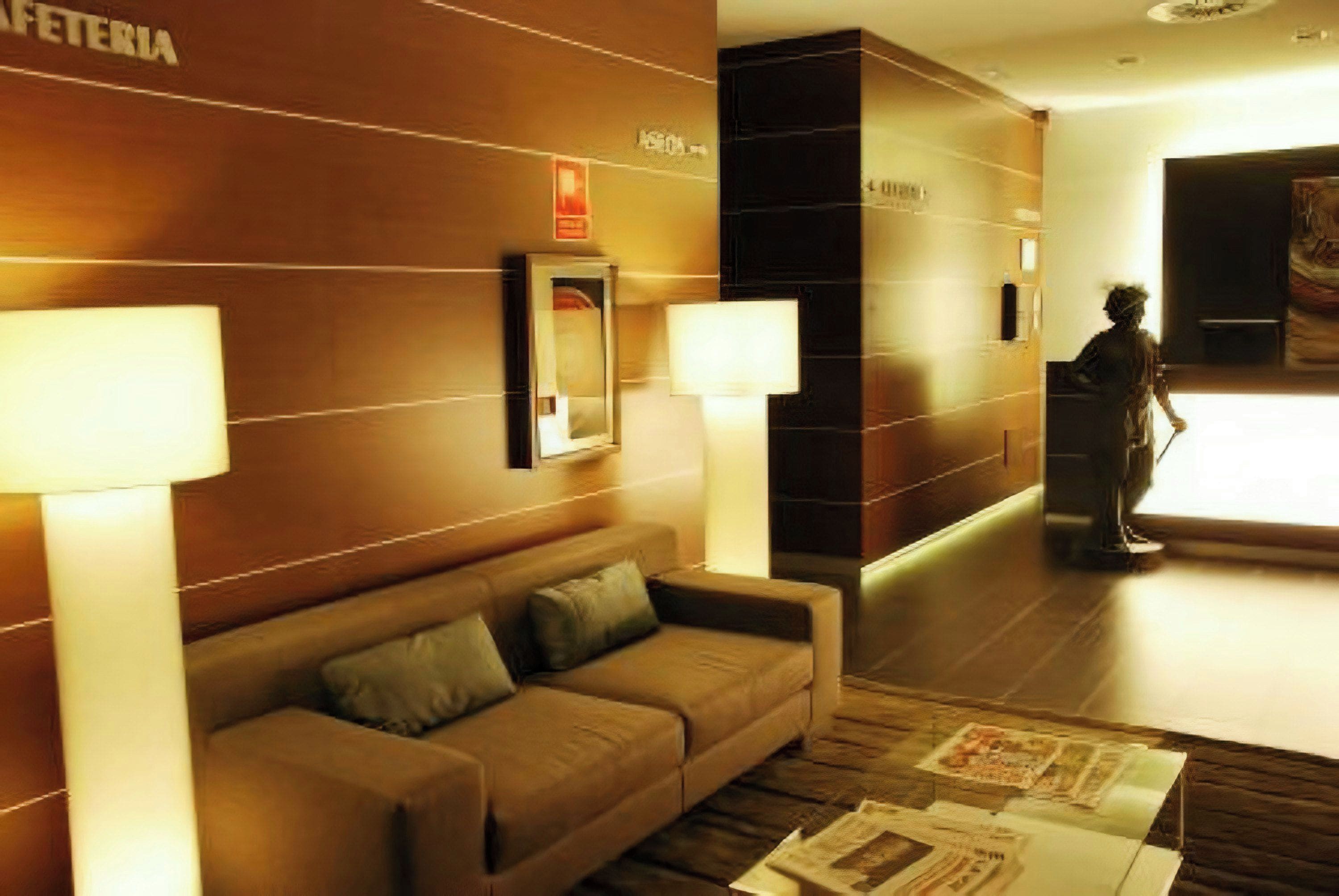 Un hall de hotel con un sofá, dos lámparas de pared, un cuadro enmarcado y una persona de pie cerca de una puerta.