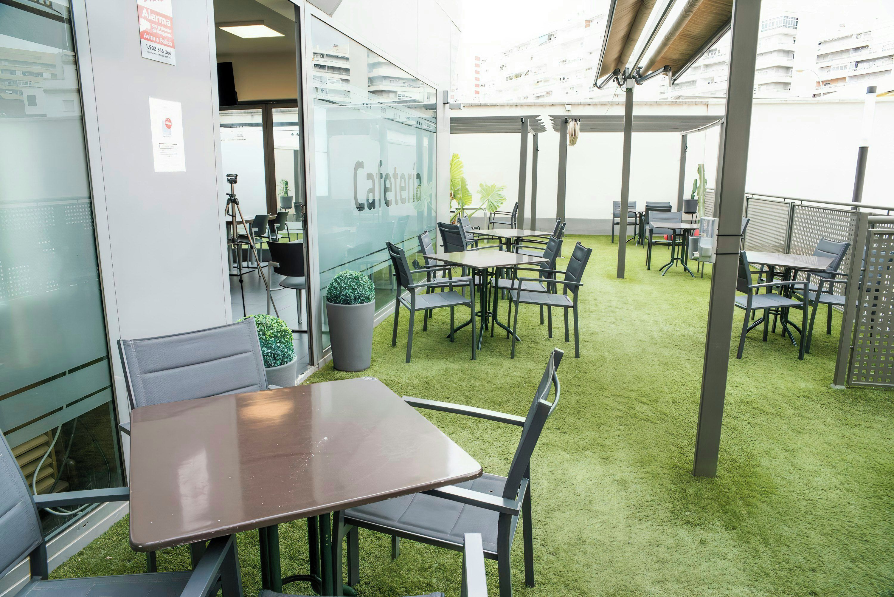 An outdoor café area with tables and chairs, a green artificial turf floor, and a glass wall with the word 'Cafeteria' on it.