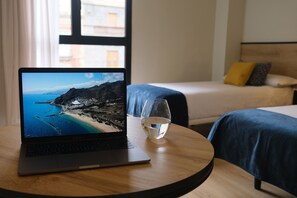 Un ordinateur portable posé sur une table ronde, à côté d’un verre d’eau, un lit avec une literie bleue et une fenêtre donnant sur un paysage côtier.