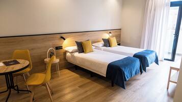 A hotel room with two beds, a small table, a chair, and a window with curtains.